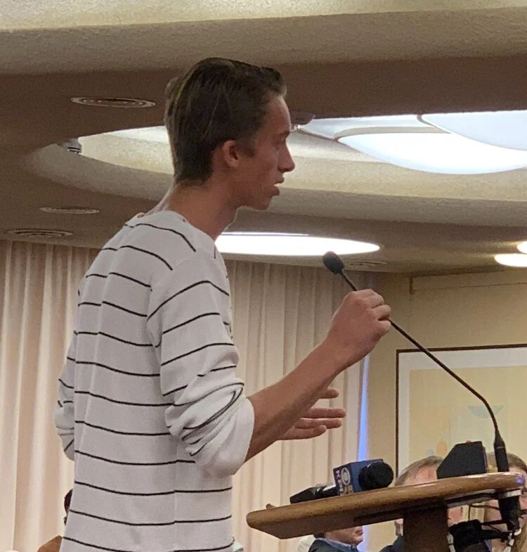 Young man speaking at a podium with microphones in a conference room.