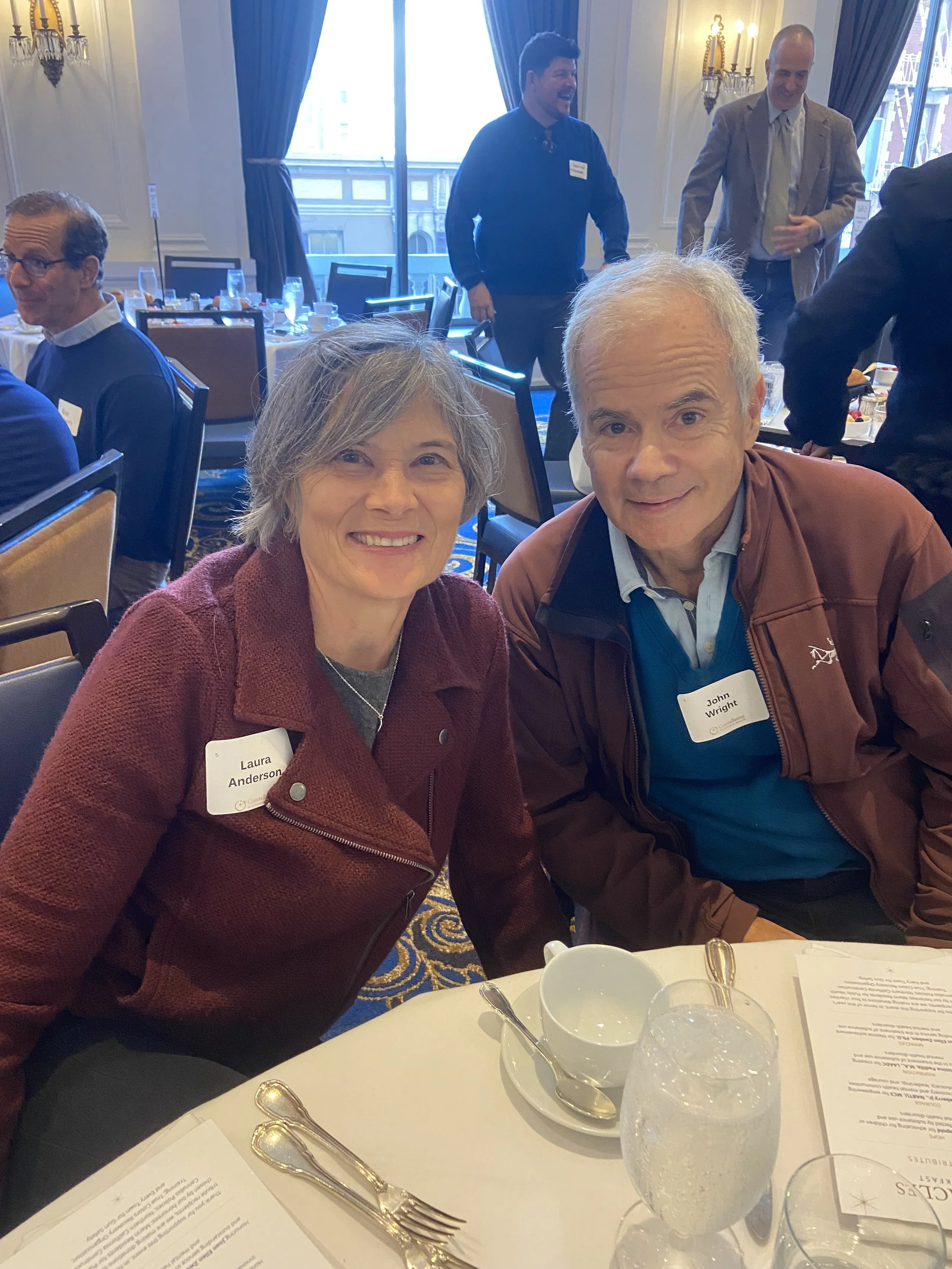 Two smiling people, a woman with gray hair wearing a maroon jacket and a man with white hair wearing a brown jacket, sitting at a table during a formal event with other attendees in the background.