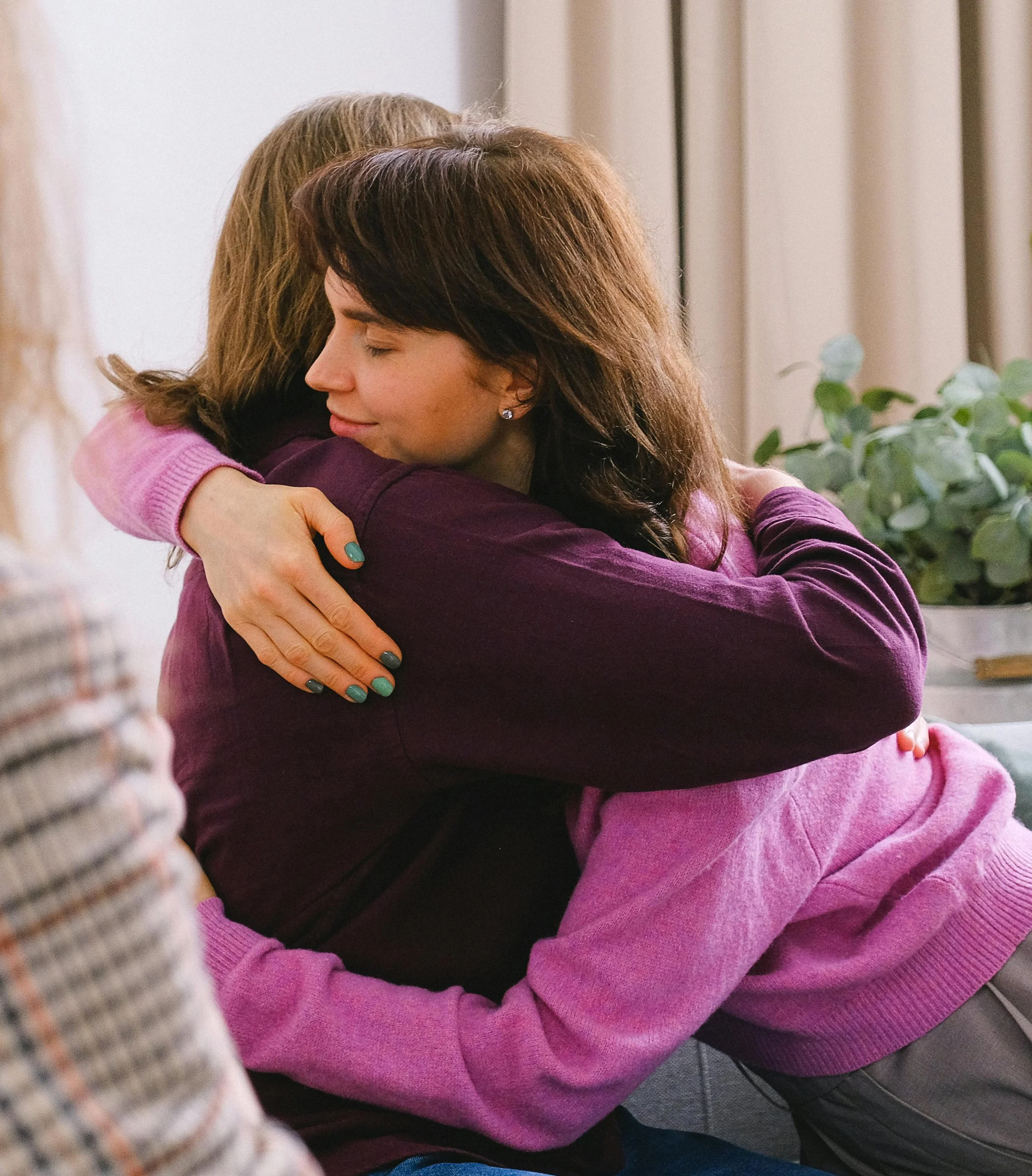Two women hugging each other warmly in a cozy setting with a plant in the background.