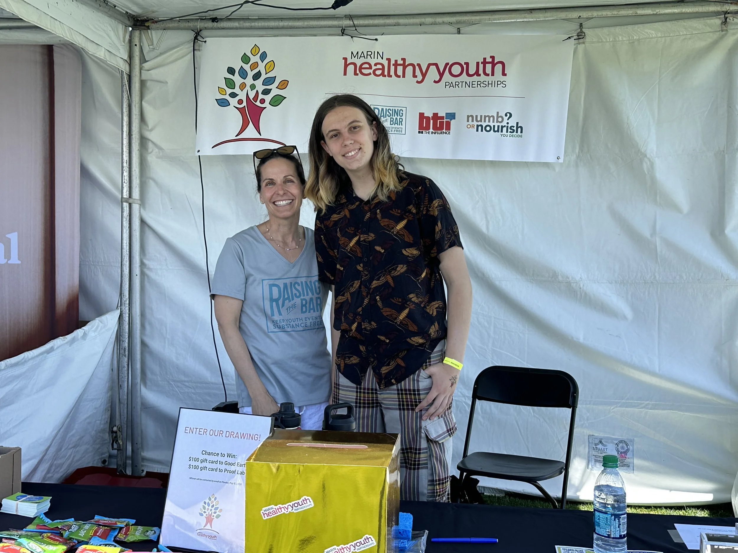 Two women standing behind a table at an outdoor event booth, smiling for the camera. The woman on the left wears a light blue t-shirt with the words "Raising the Bar" and the woman on the right wears a dark shirt with a leaf pattern. There is a banner behind them with the text "Marin healthy youth partnerships" and various logos. The table has a sign about a drawing contest, several small items, a water bottle, and a pen.