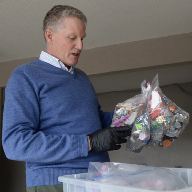 Man wearing a blue sweater and black gloves sorting through a clear plastic bag filled with empty pill bottles, with more bottles in a container below.