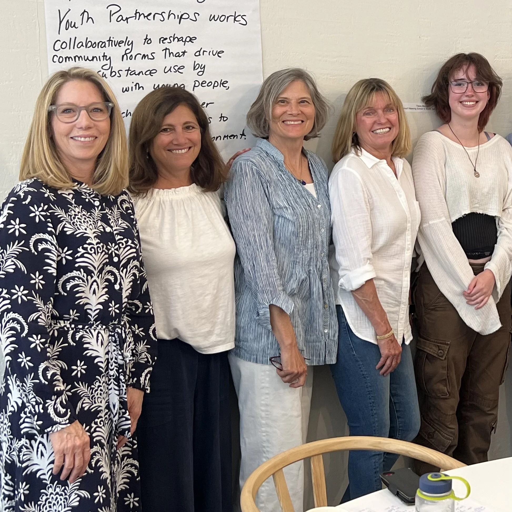 Group of five women standing together indoors, smiling for the camera, with a handwritten poster on the wall behind them and a table with a water bottle and electronic device in front of them.