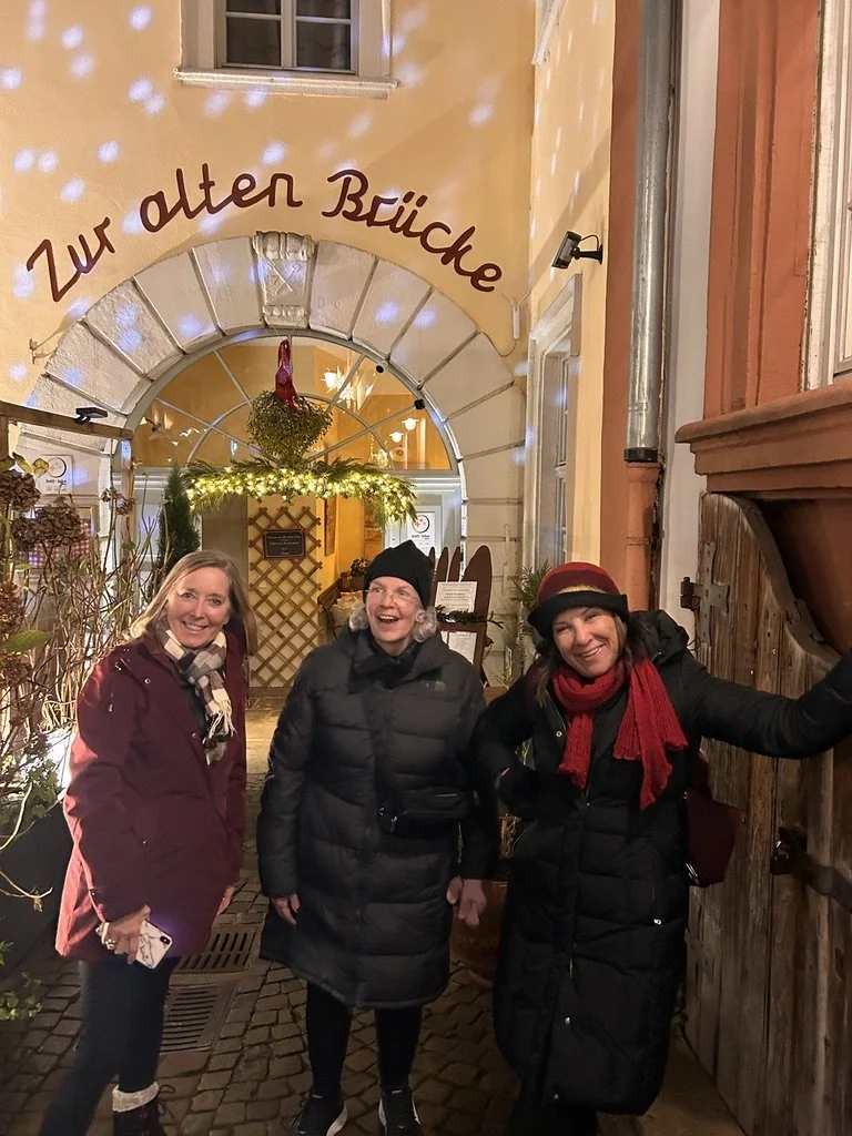 Three travelers laughing outside a lit doorway on a cobblestone street at night