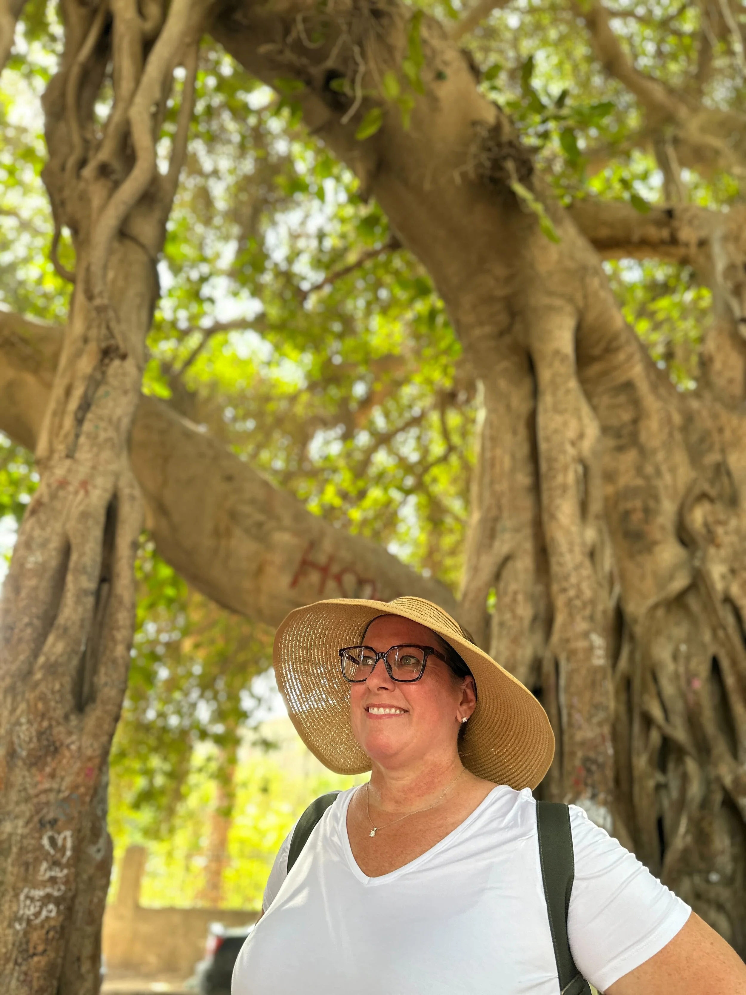 Woman with glasses and a wide-brimmed hat smiling outdoors in front of a large, old tree with twisting branches and green foliage.