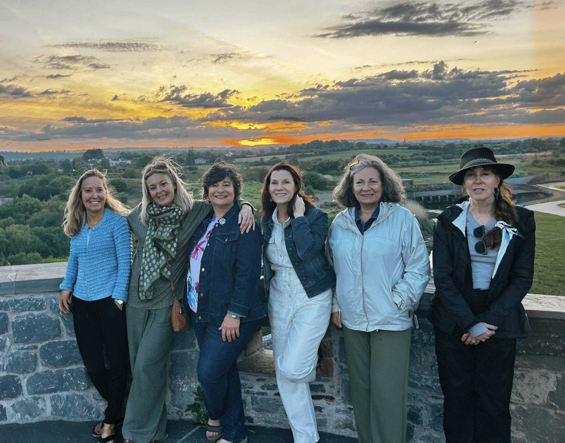 Six women standing together outdoors on a stone wall at sunset with a scenic landscape of fields and a river in the background.