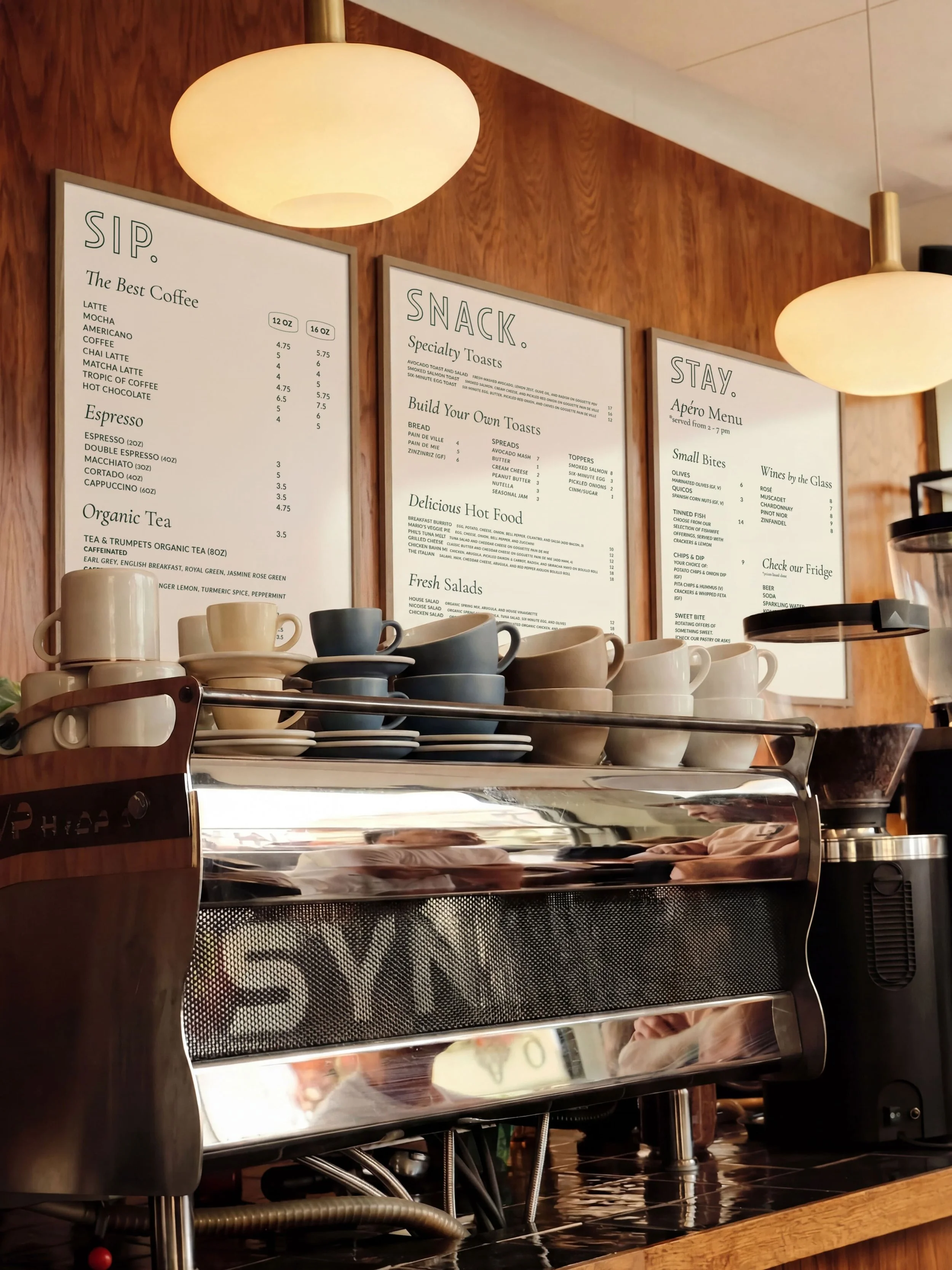 Close-up of a coffee shop menu board with various drink options and prices, with cups and a coffee machine in the foreground.