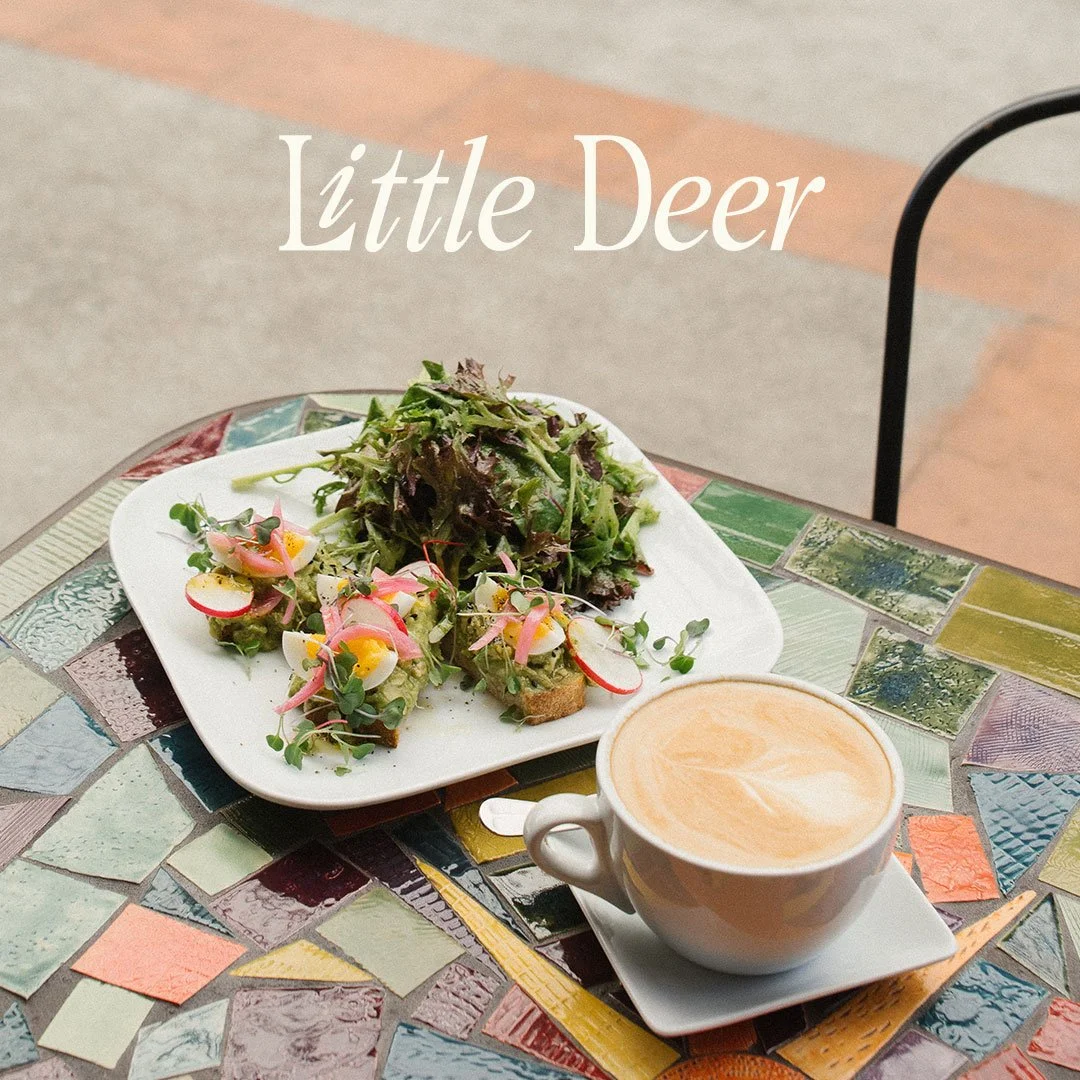 A table with a colorful tiled surface holds a white plate of salad greens with radishes, eggs, and microgreens, a cup of latte, and the words "Little Deer" over the image.