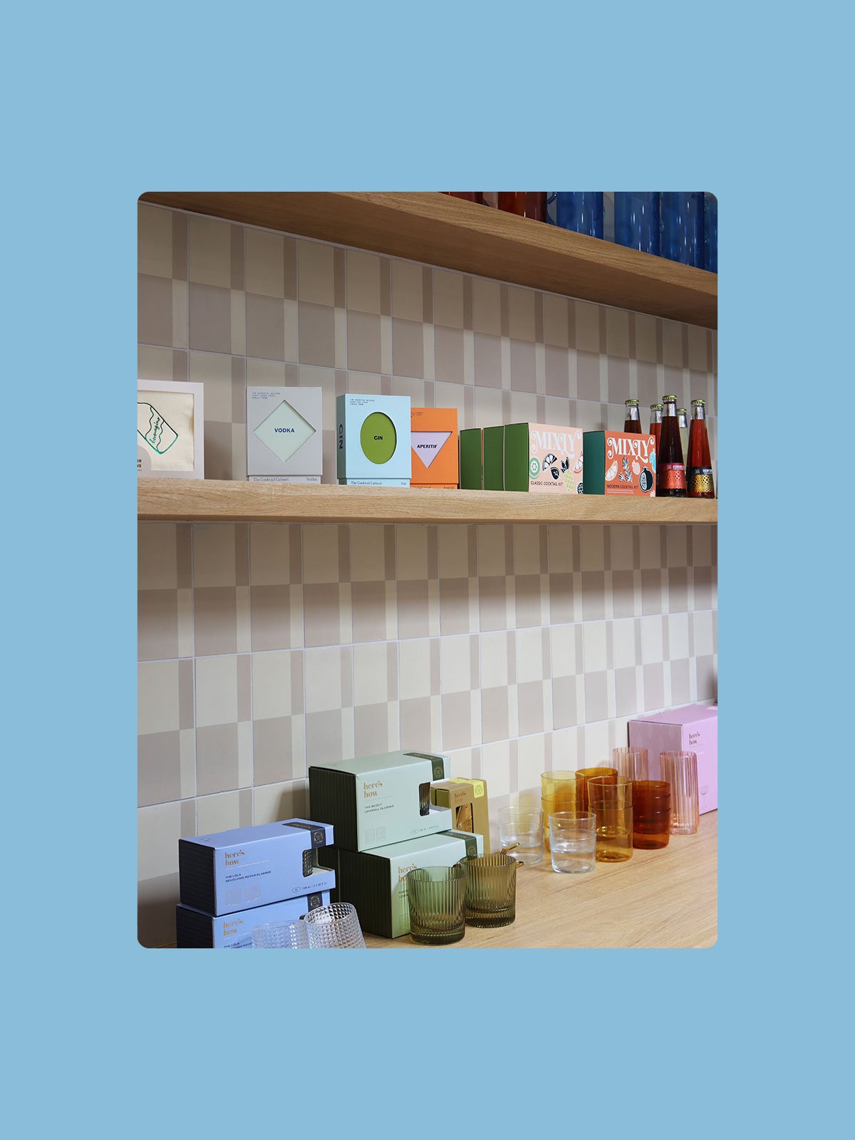 Display of colorful glasses and boxed tableware on a wooden shelf and table against a tiled wall.