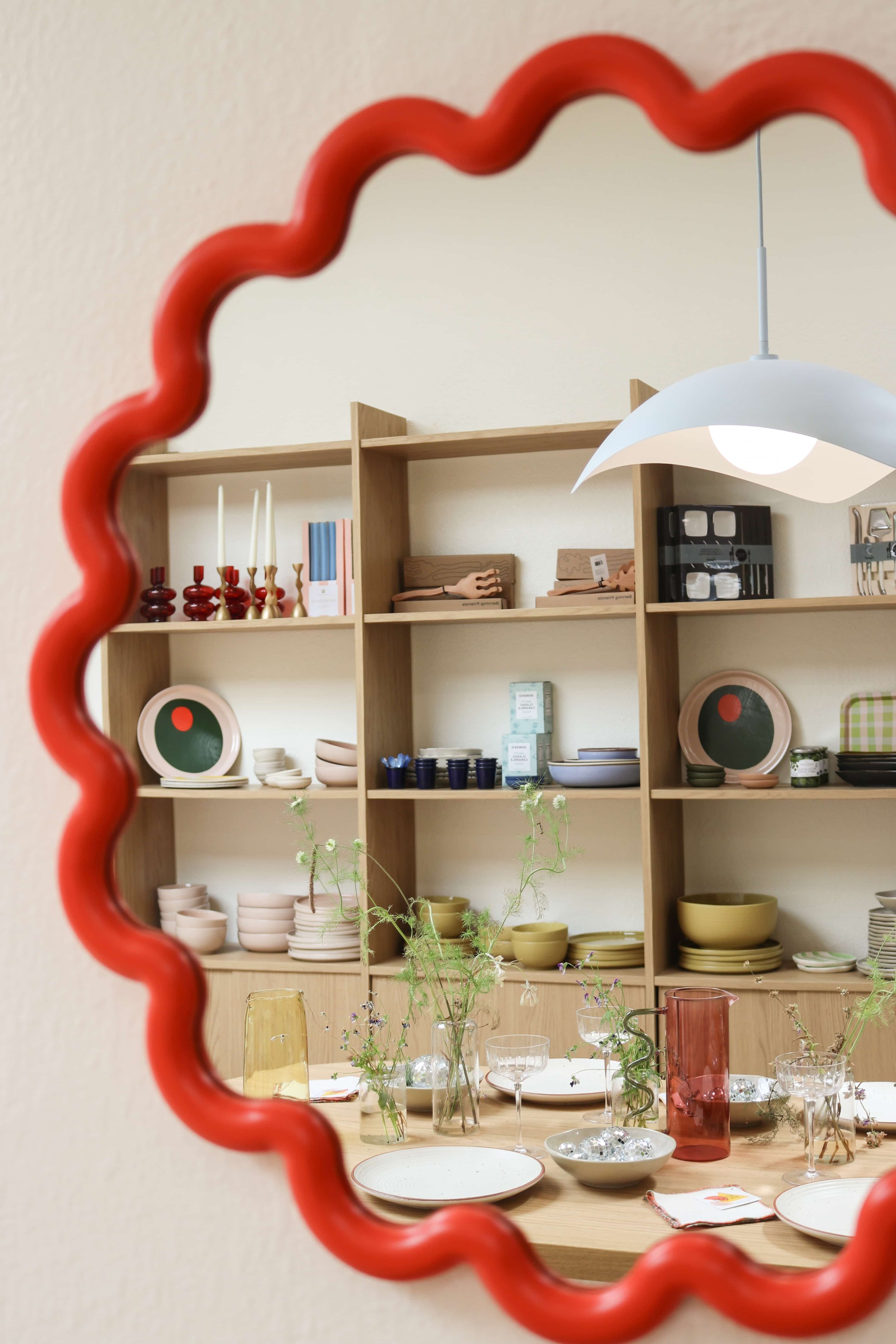 A mirror with a wavy red border reflecting a wooden shelf with candles, books, and kitchenware, and a dining table with glasses and vases with flowers.