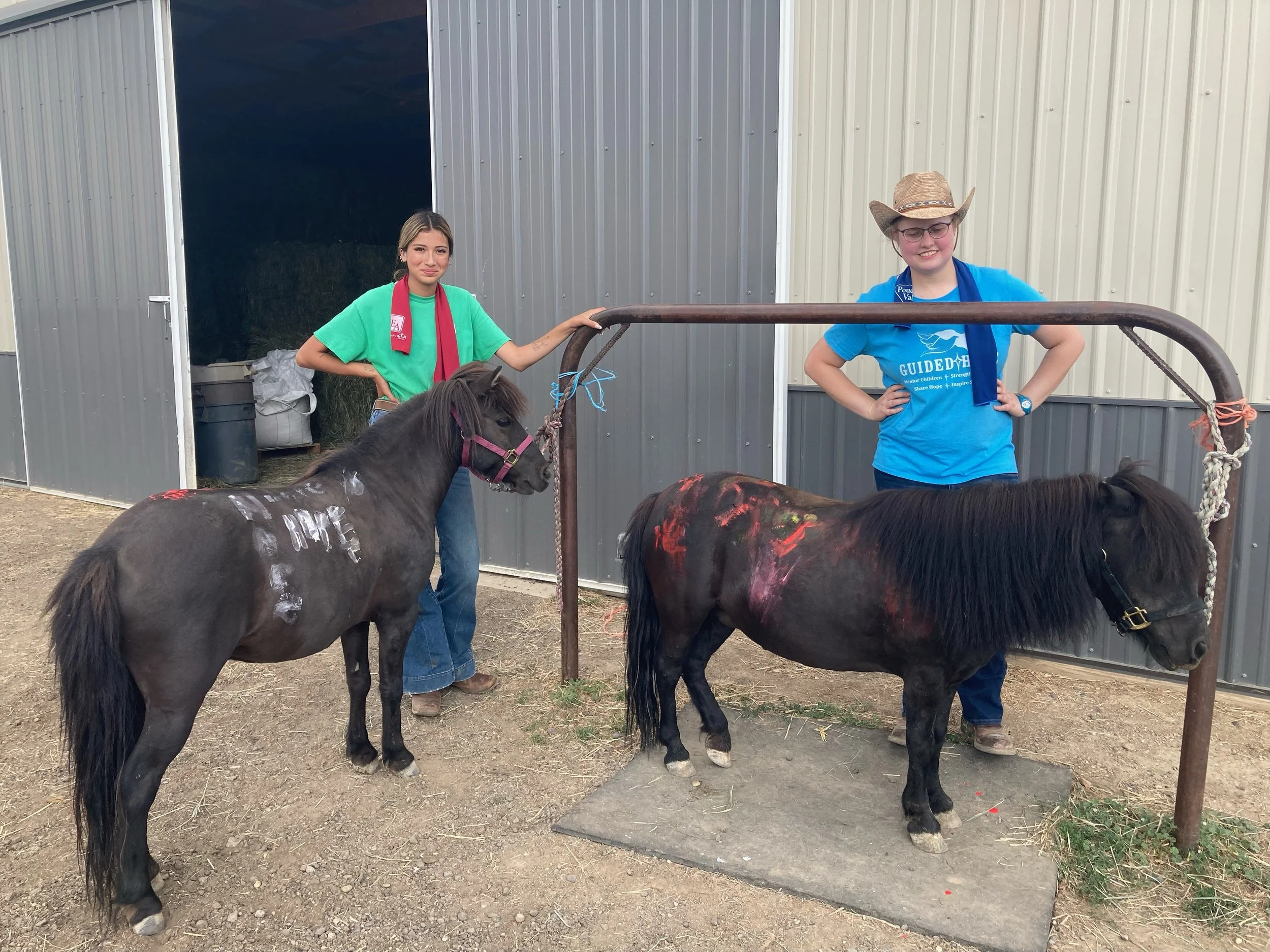 Two people stand alongside two ponies with painted designs, in front of a barn with an open door.