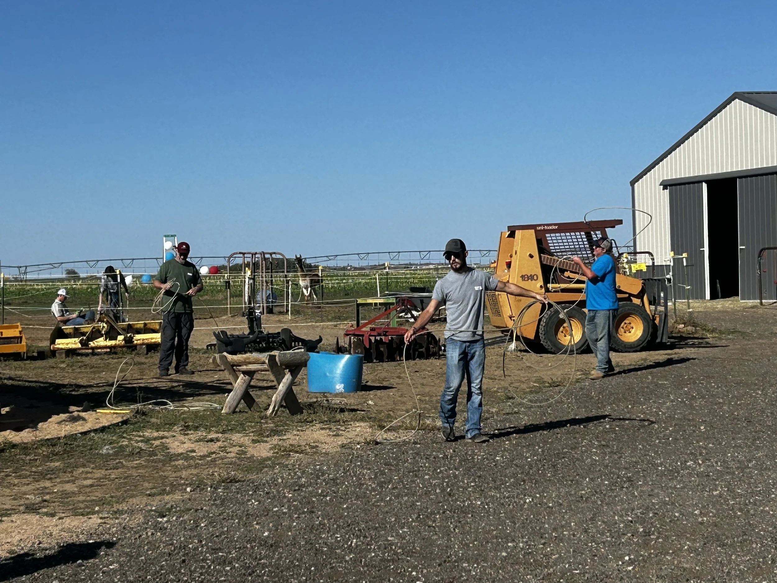 Men practicing roping skills outdoors on a farm near farming equipment and a barn.