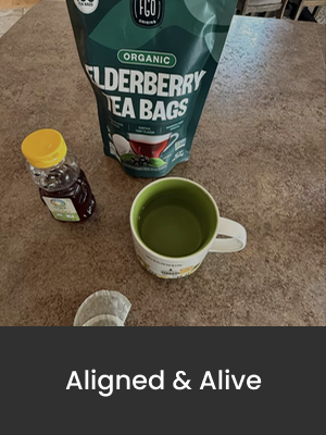 A bag of organic elderberry tea bags, a bottle of elderberry syrup, and a mug filled with green tea on a countertop.