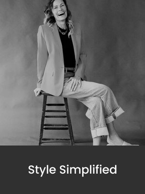 A woman with wavy hair wearing a blazer, black top, and wide-leg jeans, laughing while sitting on a wooden stool against a plain background.