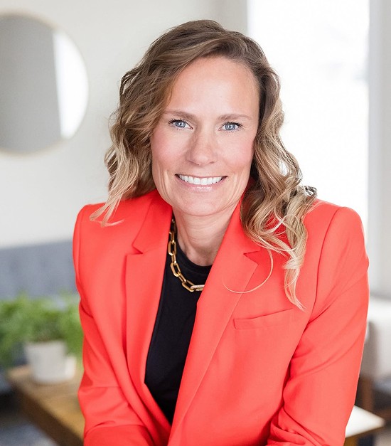 A woman with blonde, wavy hair, smiling, wearing a coral blazer over a black top, and a gold chain necklace, sitting indoors with a blurred background.