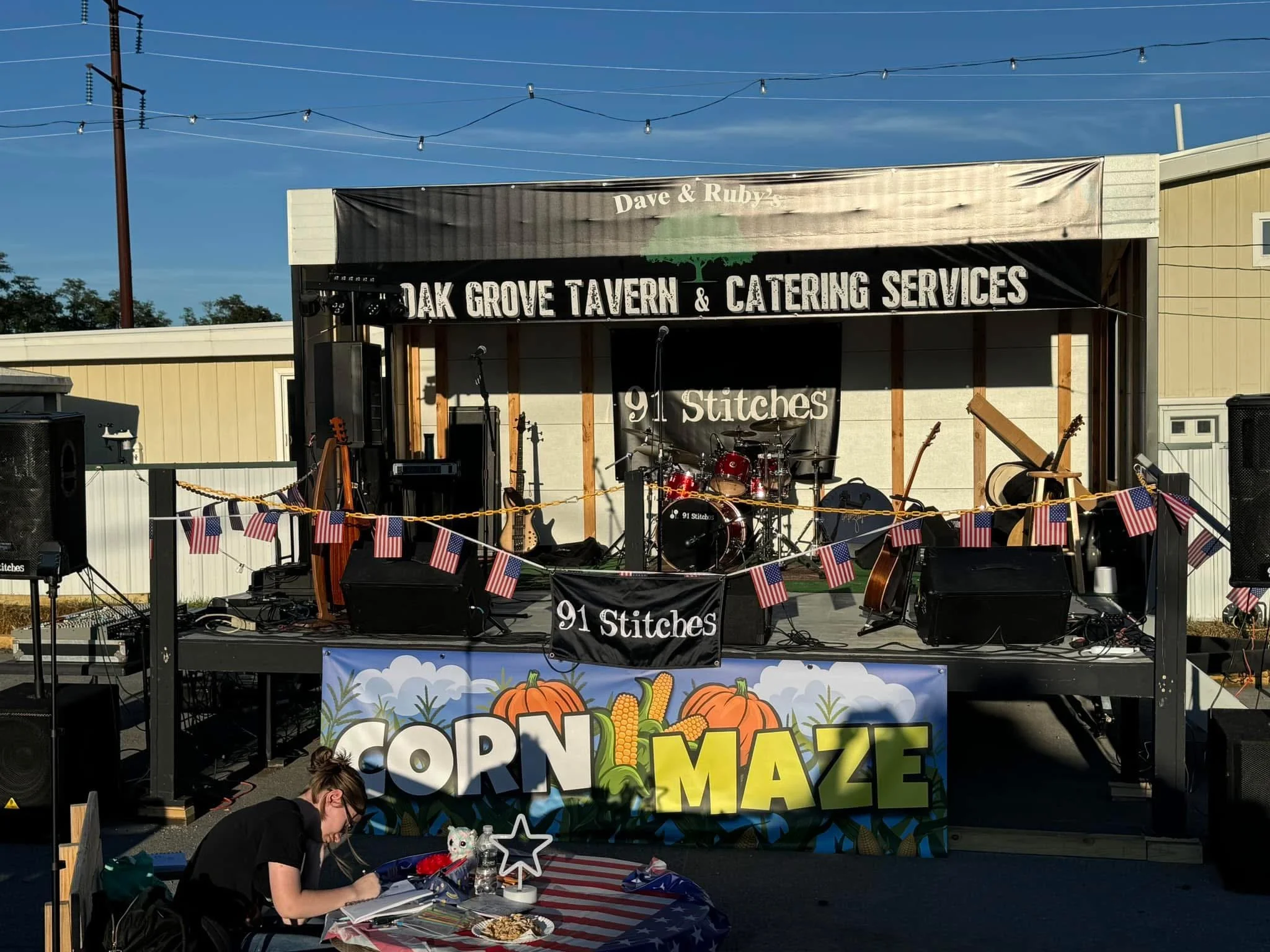 Stage with musical instruments at an outdoor event, decorated with a corn maze banner, American flags, and a person sitting at a table in front of it.