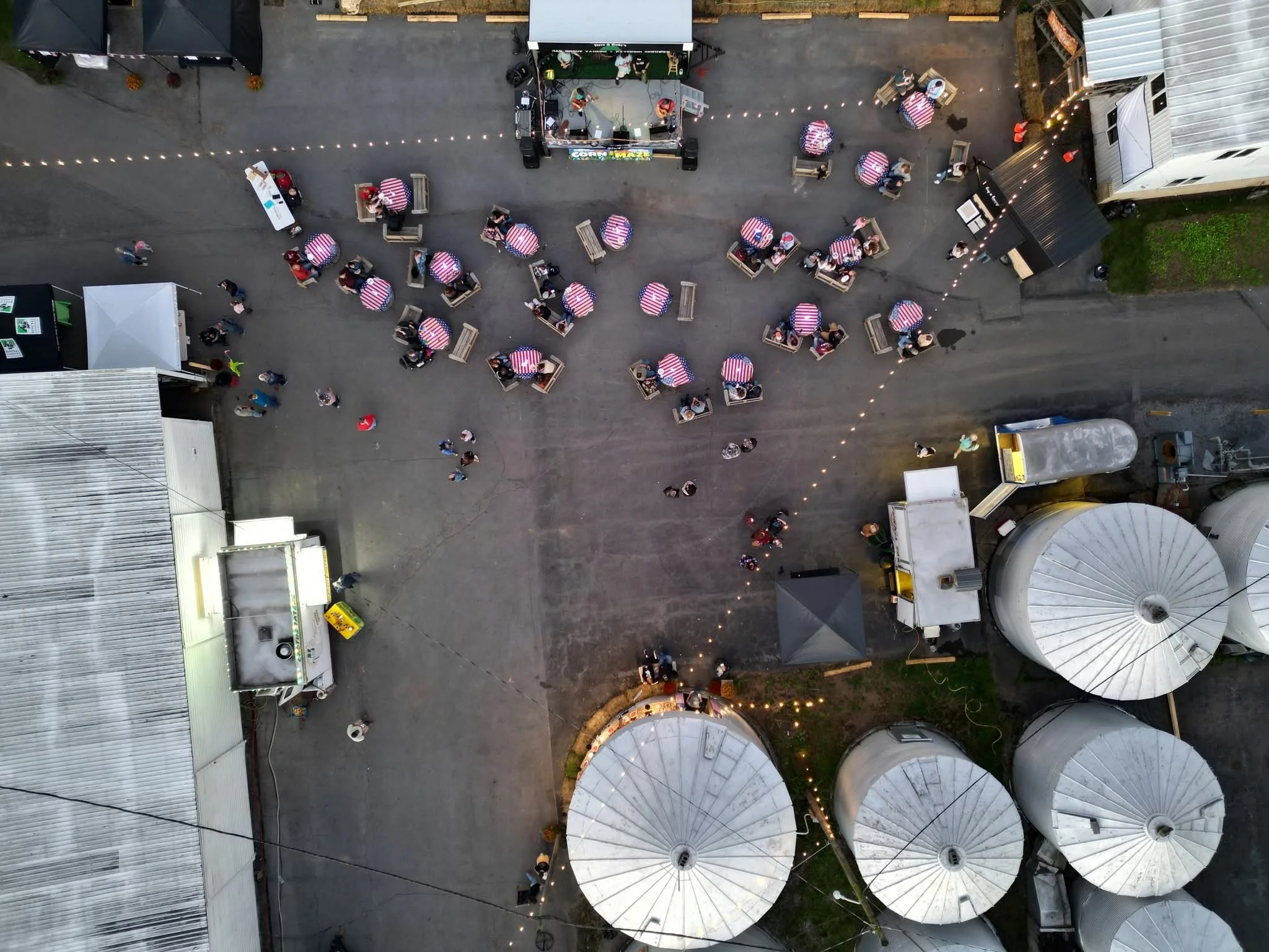 An aerial view of an outdoor gathering with numerous tables covered with red, white, and blue tablecloths, surrounded by people, string lights, food trucks, and structures with metal roofs.