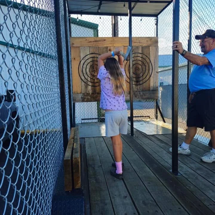 A girl in a purple shirt and shorts throws an axe at a target, supervised by a man in a blue shirt and cap at an outdoor axe throwing range.