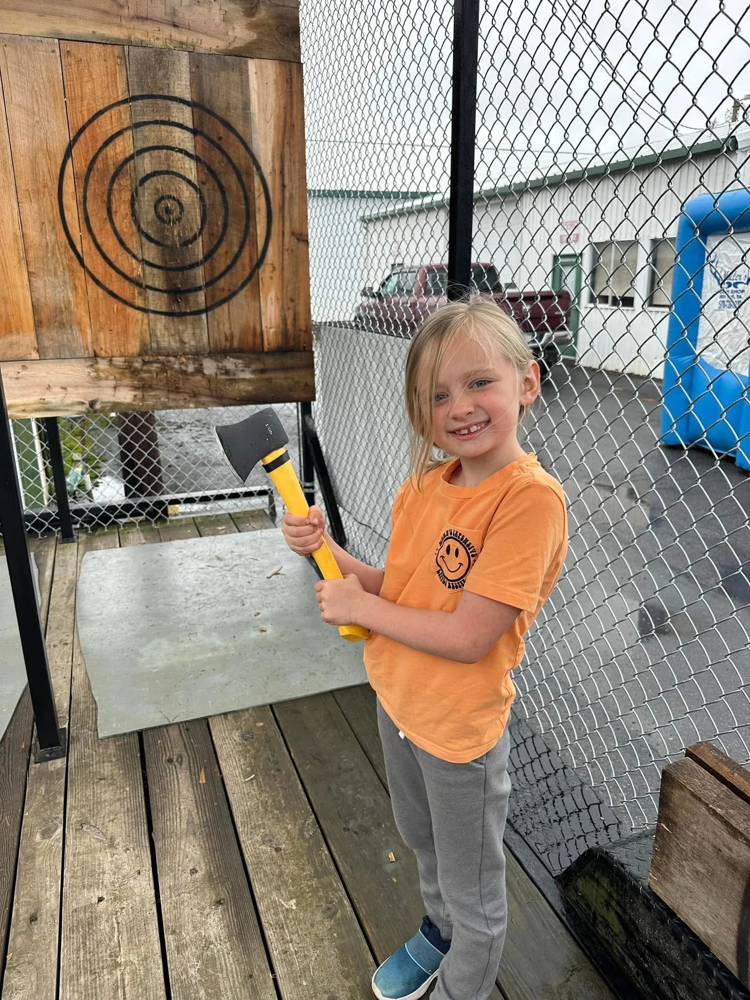 Young girl with blonde hair, smiling, wearing an orange t-shirt with a smiley face and grey pants, holding a small axe, ice skate shoes, standing outside on a wooden deck near a target on a wooden board, with a chain-link fence and parked cars in the