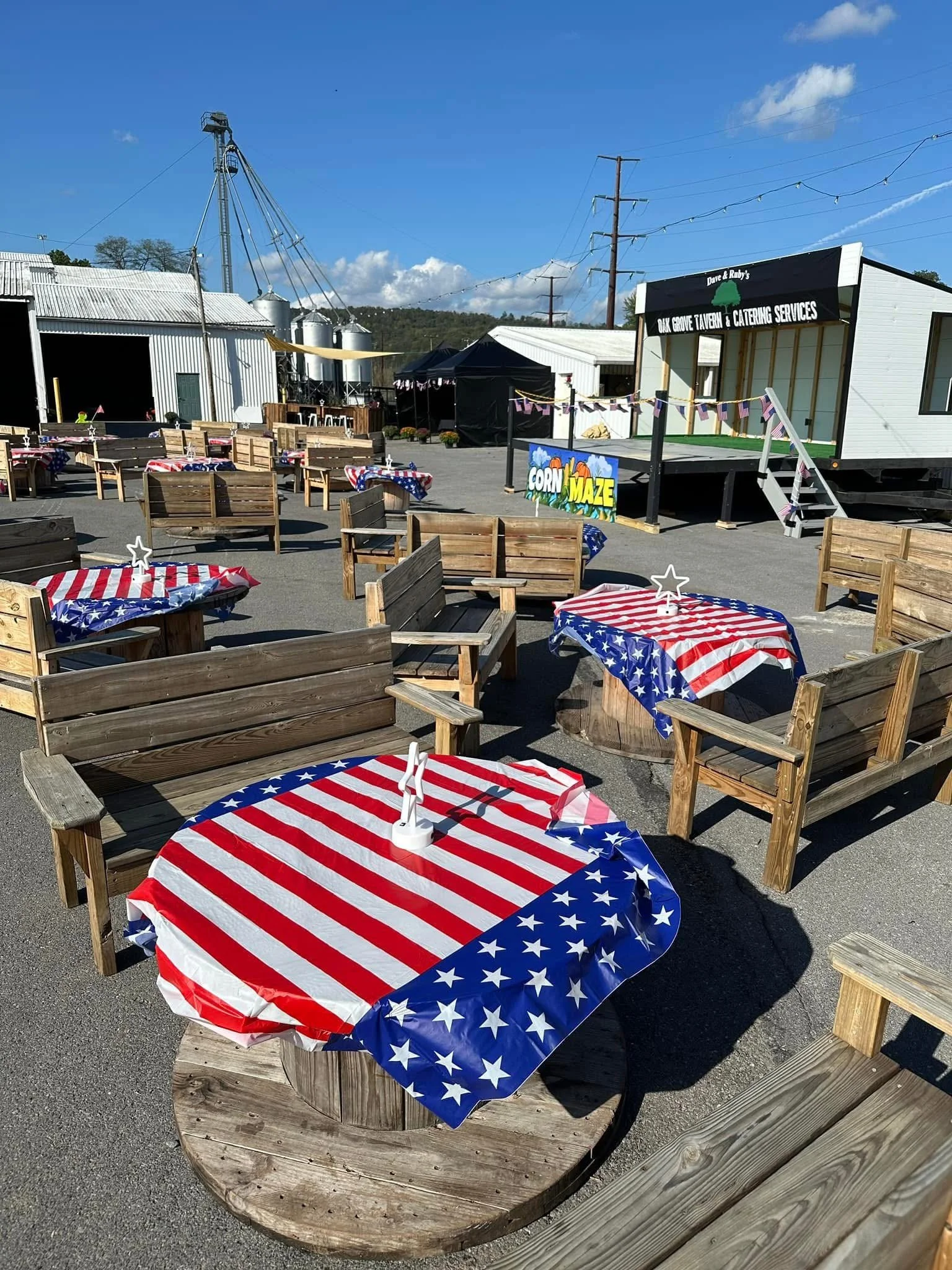 Outdoor patio area with tables covered in red, white, and blue American flag tablecloths, wooden benches and chairs, and a stage with a sign for Dave & Ruby's Oak Grove Tavern & Catering Services in the background.