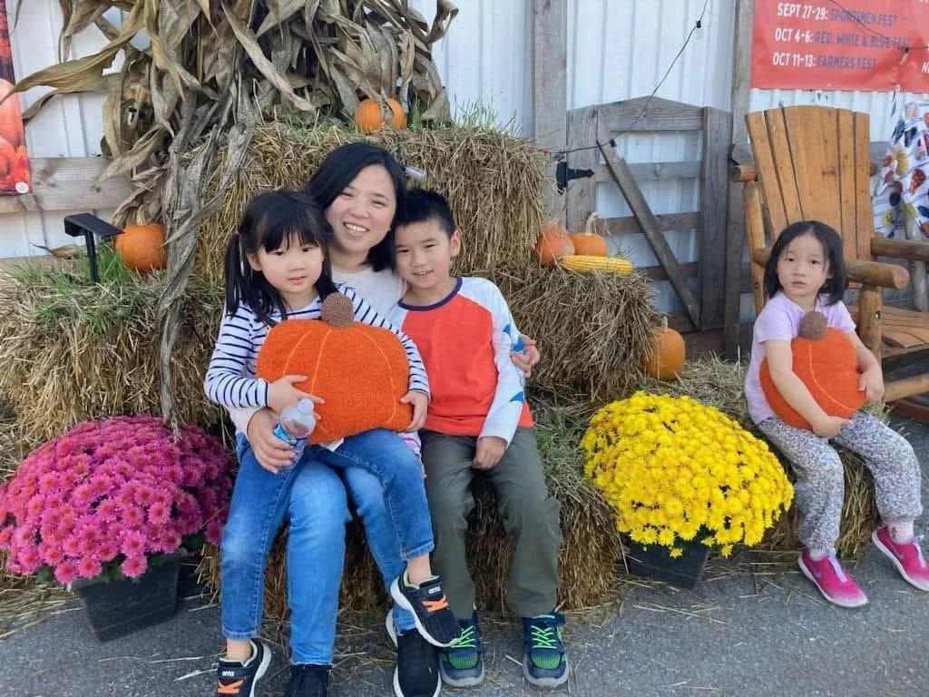 Four children and a woman sitting on hay bales surrounded by pumpkins and flowers at a fall festival.