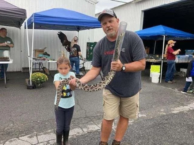 Little girl and man holding a large snake at an outdoor event with tents and people in the background.