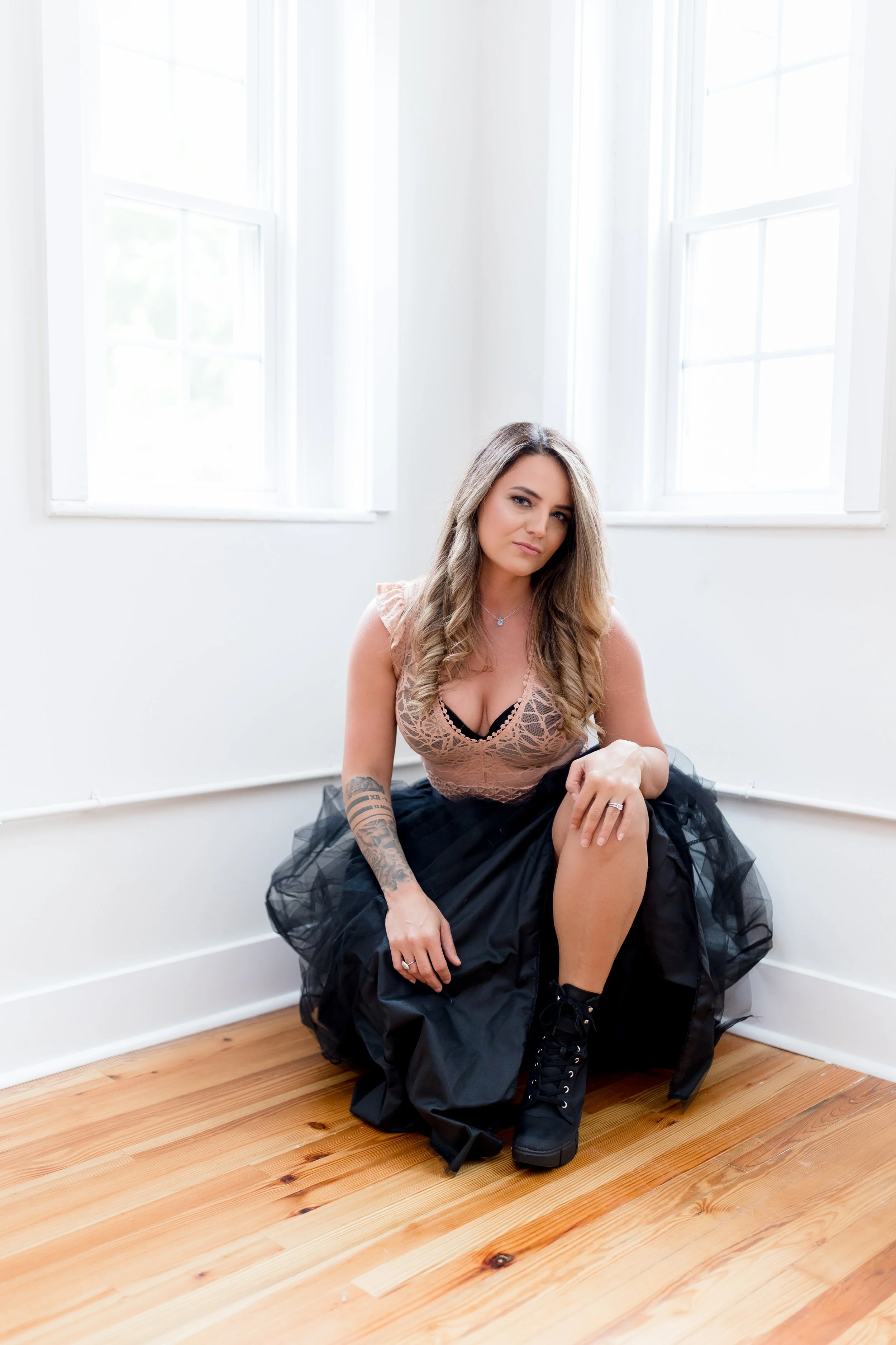 Portrait of somatic retail business coach Chrystal Rose with wavy brown hair and tattoos, with long hair and tattoos sitting on the floor in front of a white wall with two large windows, wearing a black tulle skirt, a beige lace top, and black boots.