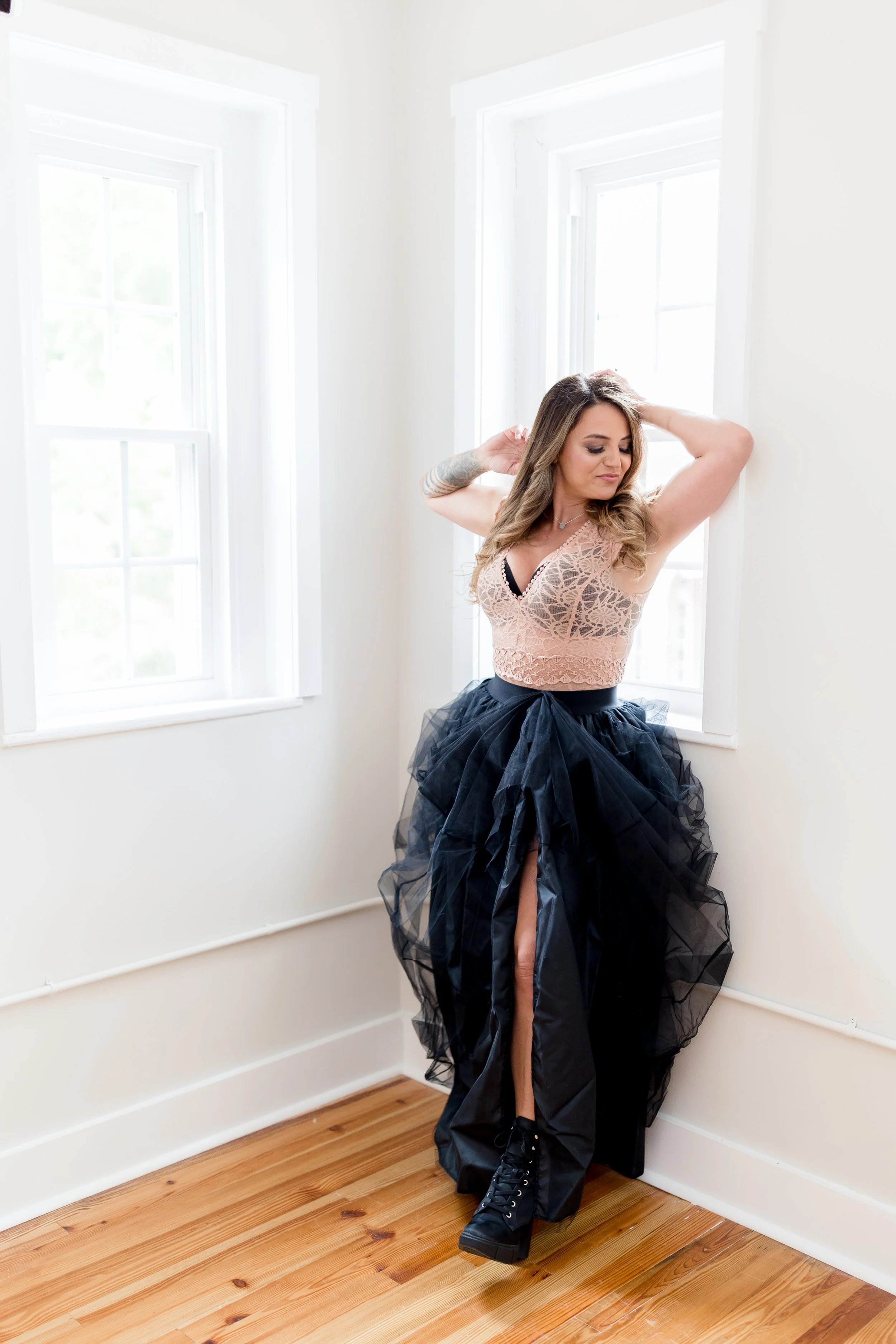 A woman with long wavy hair and tattoos on her left arm is posing by a white wall and two large windows, wearing a nude lace crop top, a long black tulle skirt featuring a high slit, and black platform shoes in a bright, sunlit room with hardwood floors.