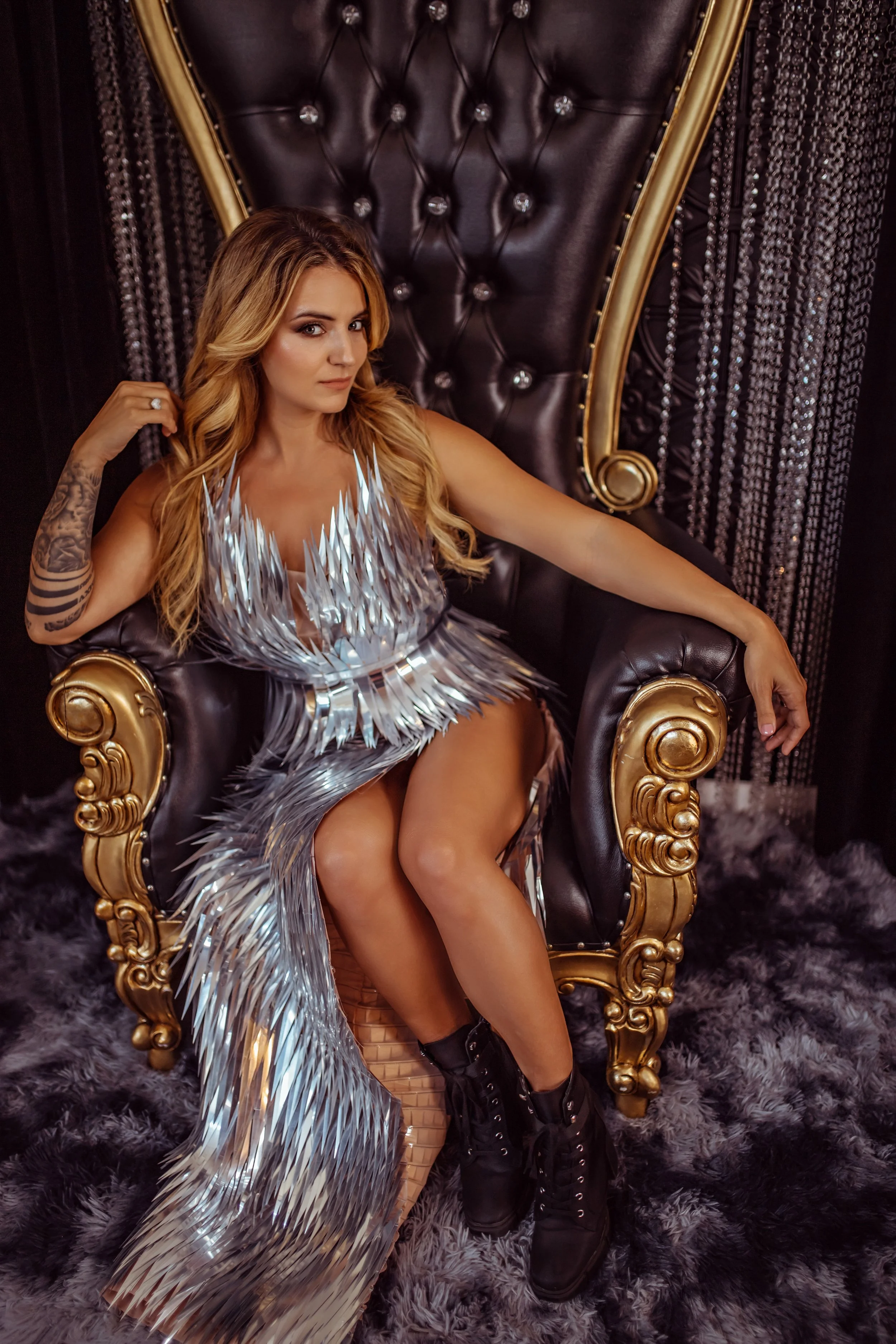 Somatic retail business coach Chrystal Rose sitting on a luxurious black and gold throne, wearing a shiny, metallic dress with geometric patterns and black combat boots. The background features beaded curtains and fuzzy carpeting.