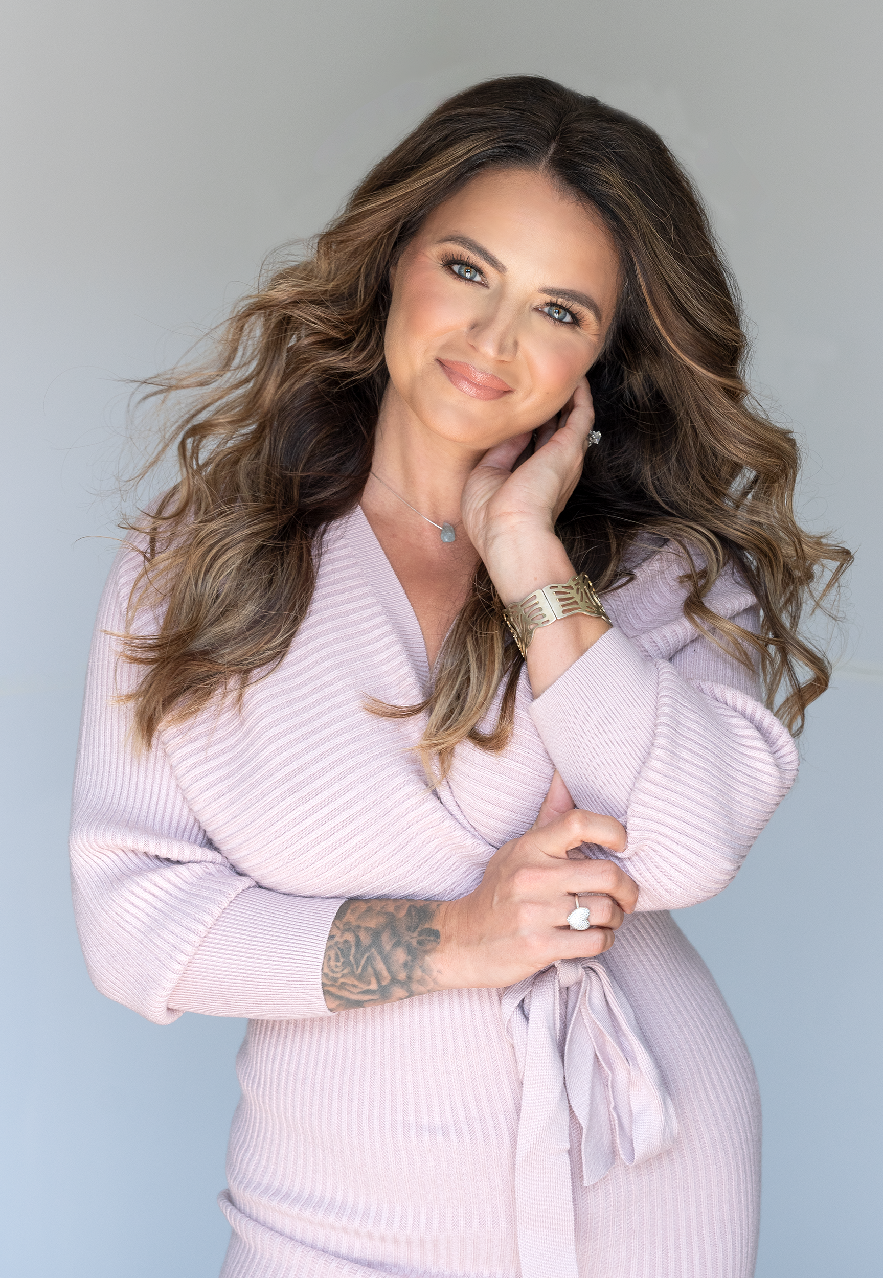 A woman with wavy brown hair and blue eyes wearing a light pink sweater, gold jewelry, and a large ring, posing with her hand near her face and a tattoo on her forearm.