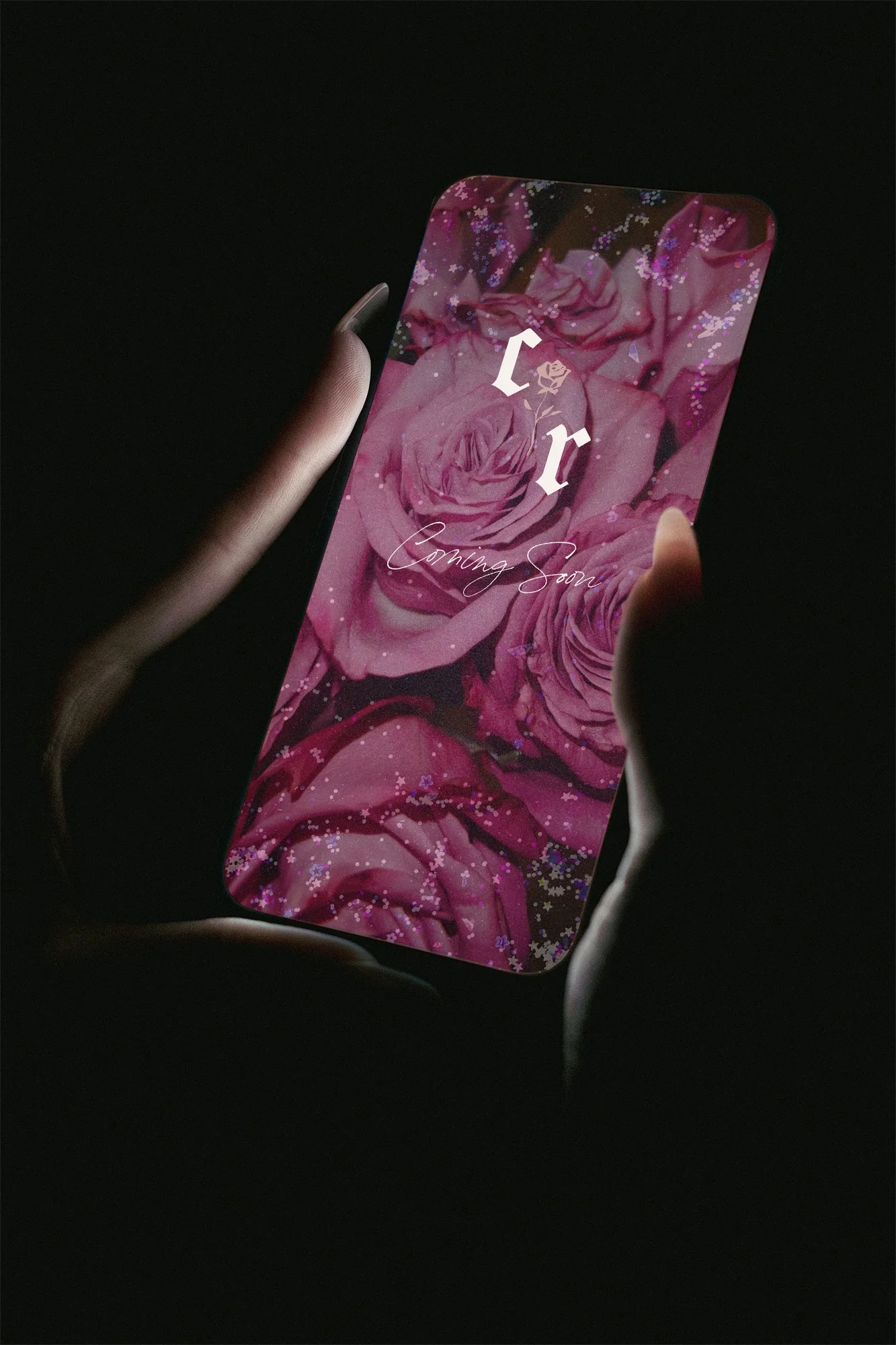 Close-up of a smartphone screen showing a floral background with pink roses and the words "Coming Soon" written in cursive, held by a person's hand against a dark background.