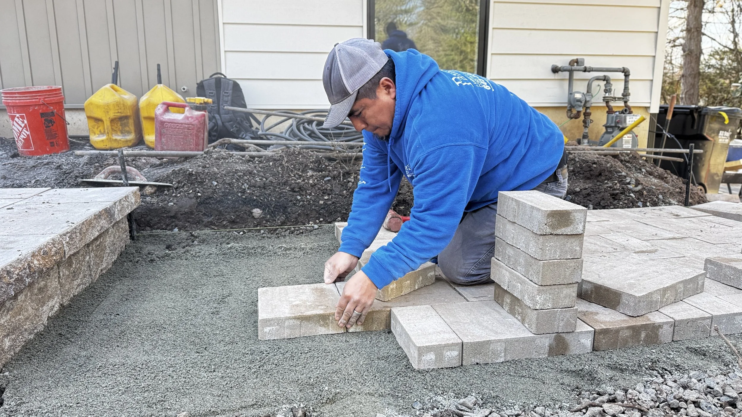 A man wearing a blue hoodie and cap is laying bricks on a concrete slab during a construction project outside a house.