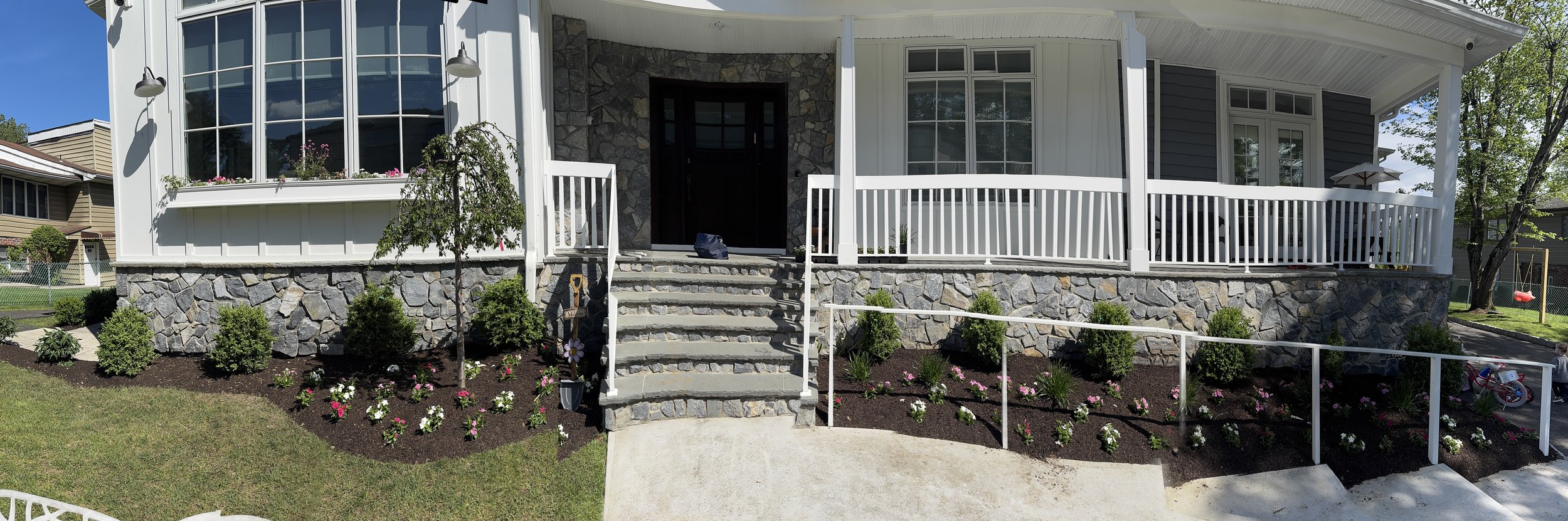 Front view of a house with a stone foundation, white railing, and steps leading to a black front door. The yard features flowering plants and bushes, with a garden in the background.