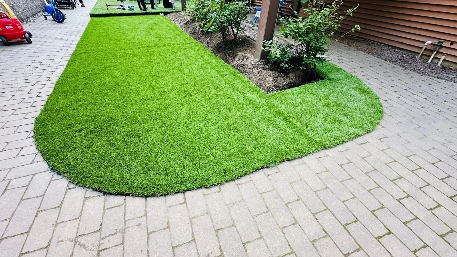 Artificial grass lawn with curved edges and a small garden bed with shrubs, bordered by a brick patio and a wooden fence.