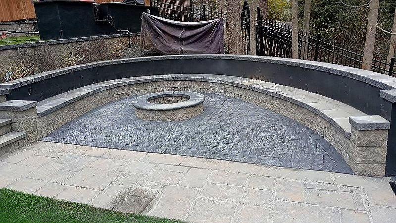 Backyard patio area with a curved stone fire pit and a stone seating wall, fenced with black metal fencing and surrounded by trees.