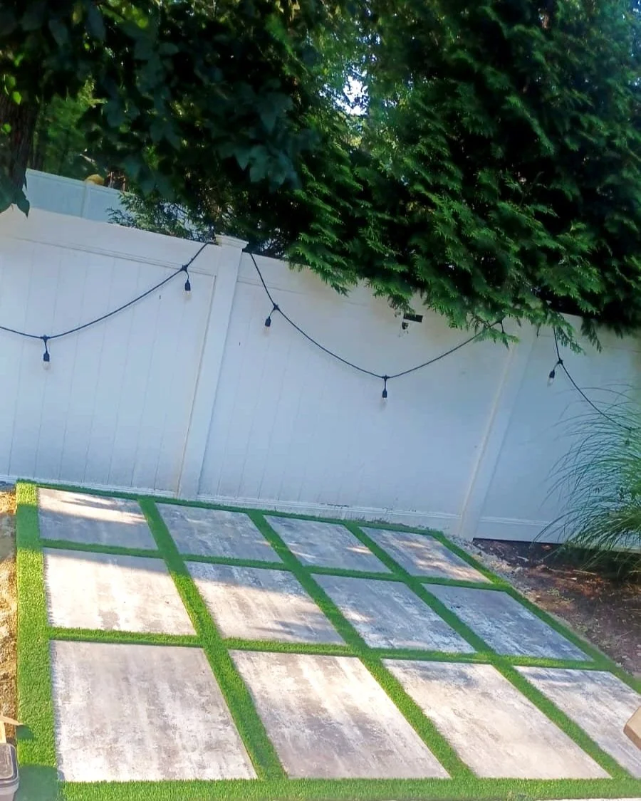 Part of a backyard patio with concrete slabs separated by green grass strips, a white fence with string lights, and leafy trees in the background