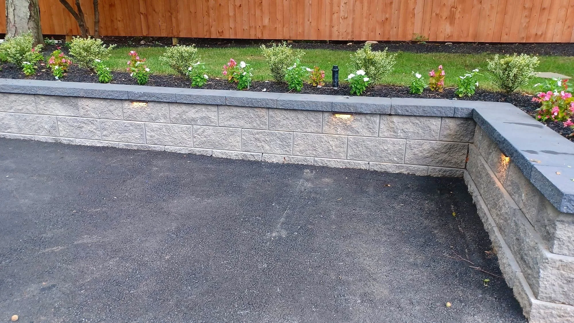 A landscaped garden bed with flowering plants and shrubs, bordered by a stone wall, with outdoor lighting beneath the plants, alongside an asphalt driveway.