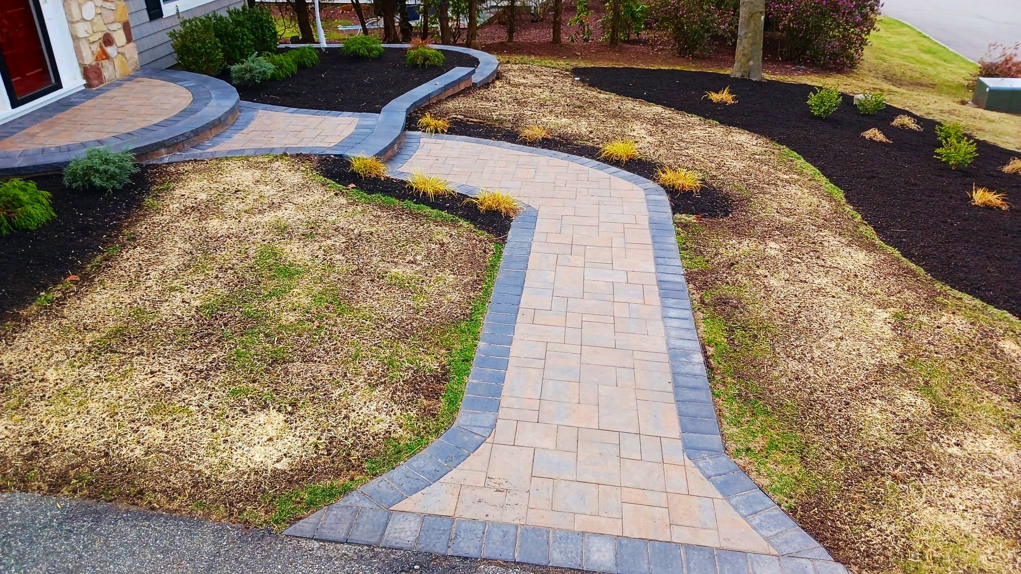 Newly paved stone walkway leading to a house entrance, bordered by black mulch and landscaped garden beds with small plants and shrubs.