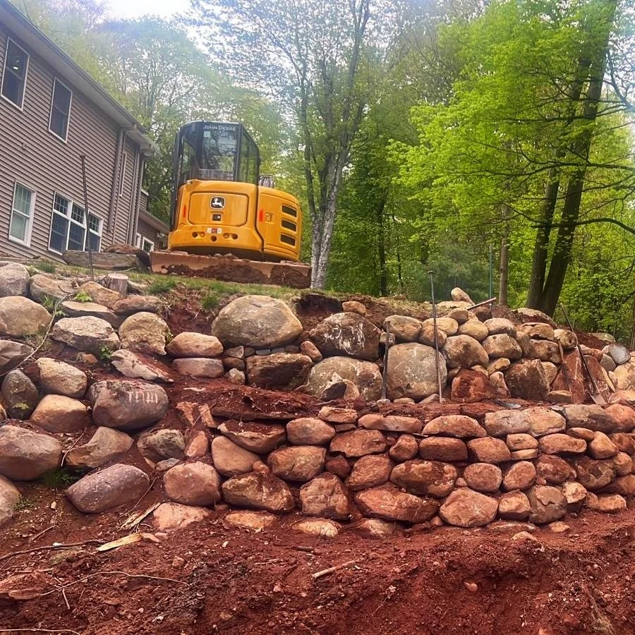 A yellow excavator working on a terraced stone wall behind a house with trees in the background.