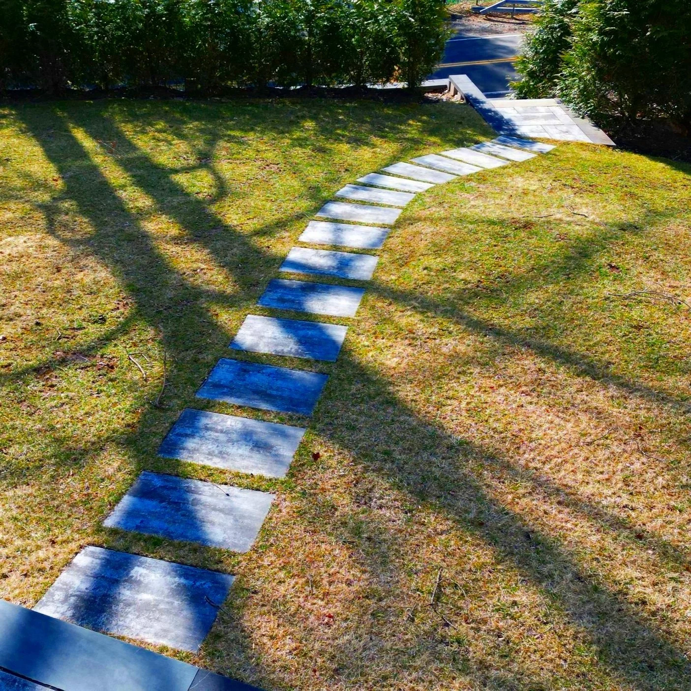 A curved stone pathway running through a well-maintained grassy yard, with long shadows cast from nearby trees on a sunny day.