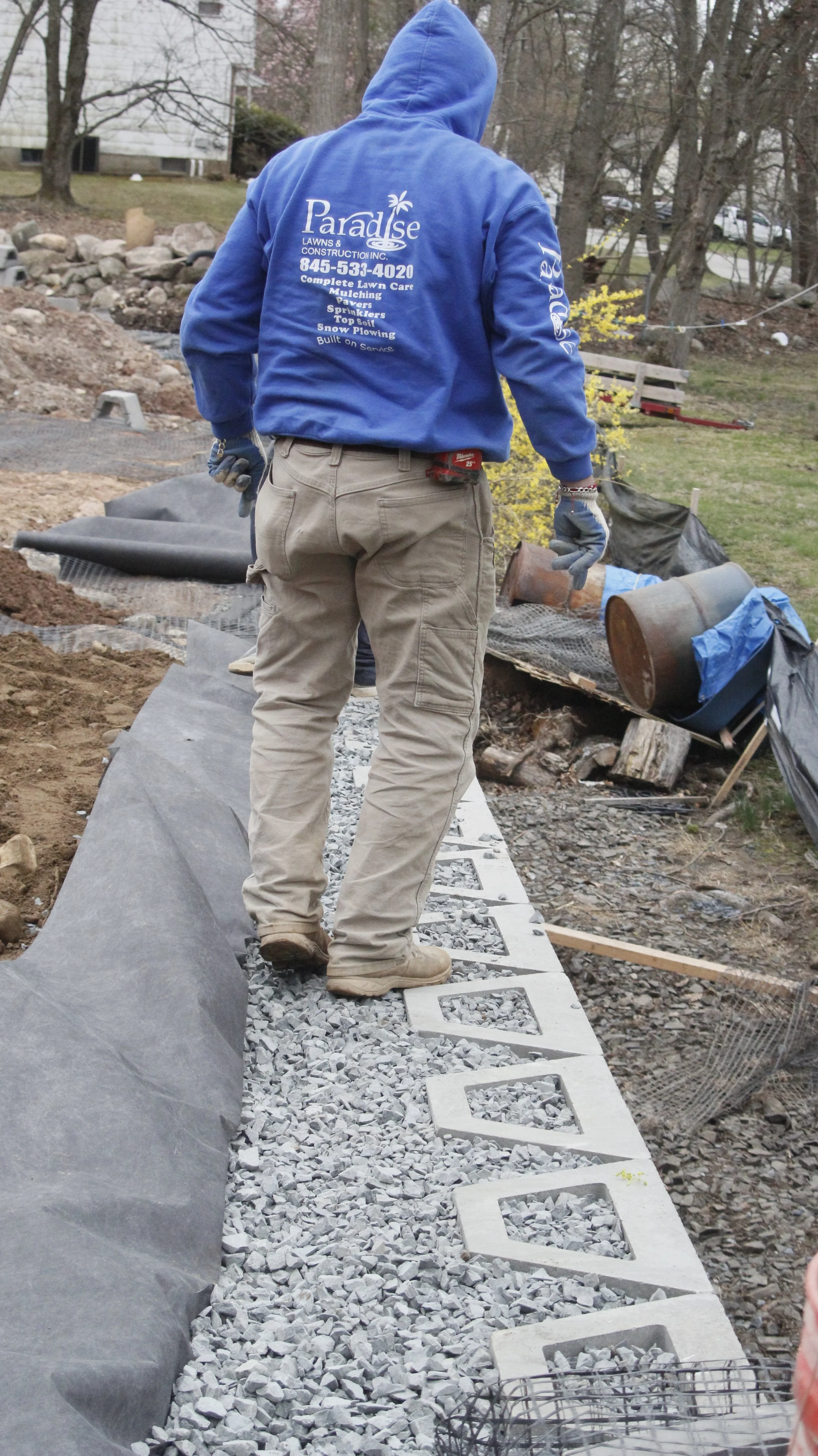 A construction worker in a blue hoodie and beige work pants installing concrete blocks on a gravel pathway in a backyard garden.