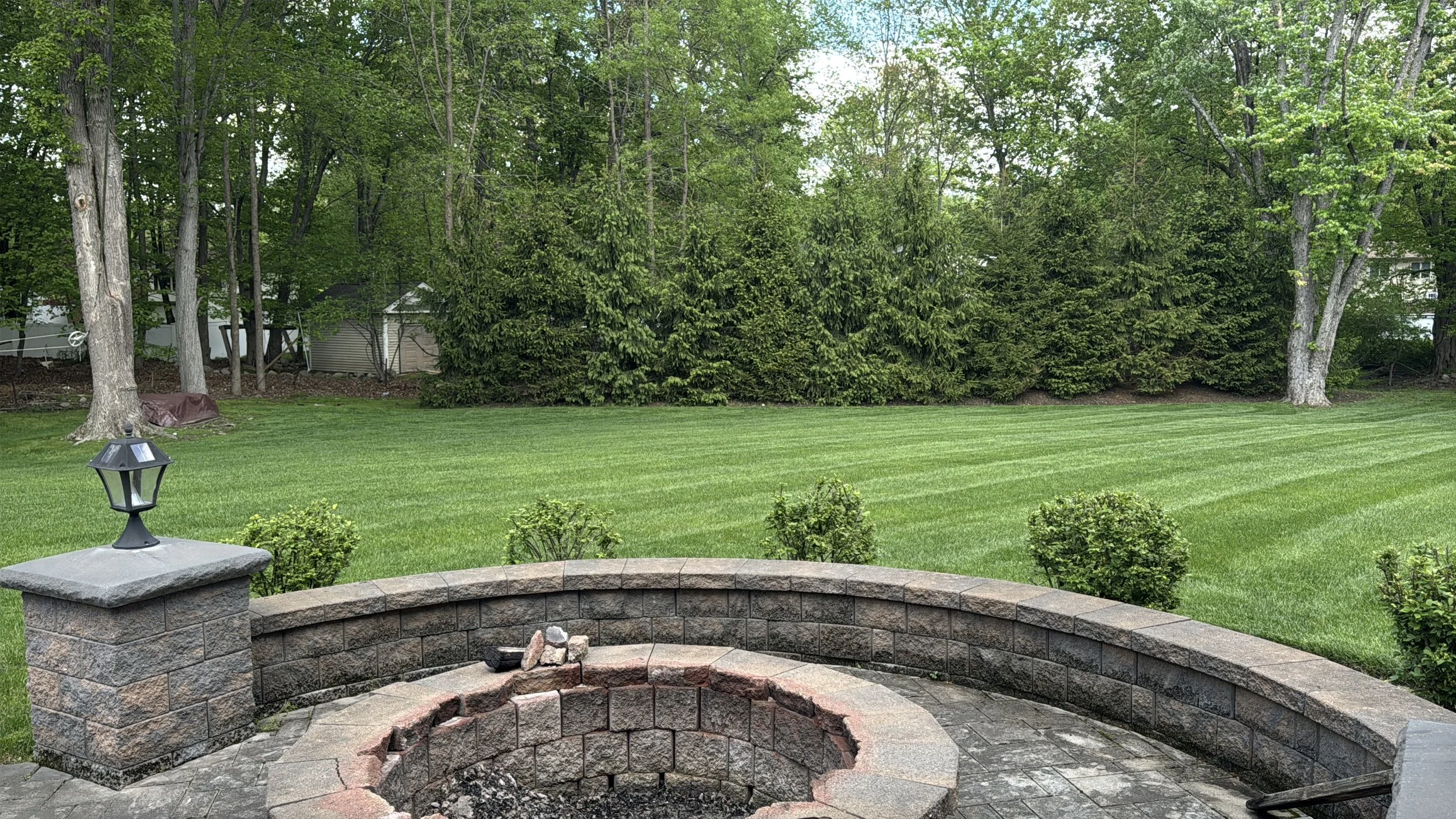 Backyard with neatly mowed grass, bushes, and tall trees, featuring a raised brick fire pit with a few small rocks on top and a black outdoor lantern on a brick pillar.