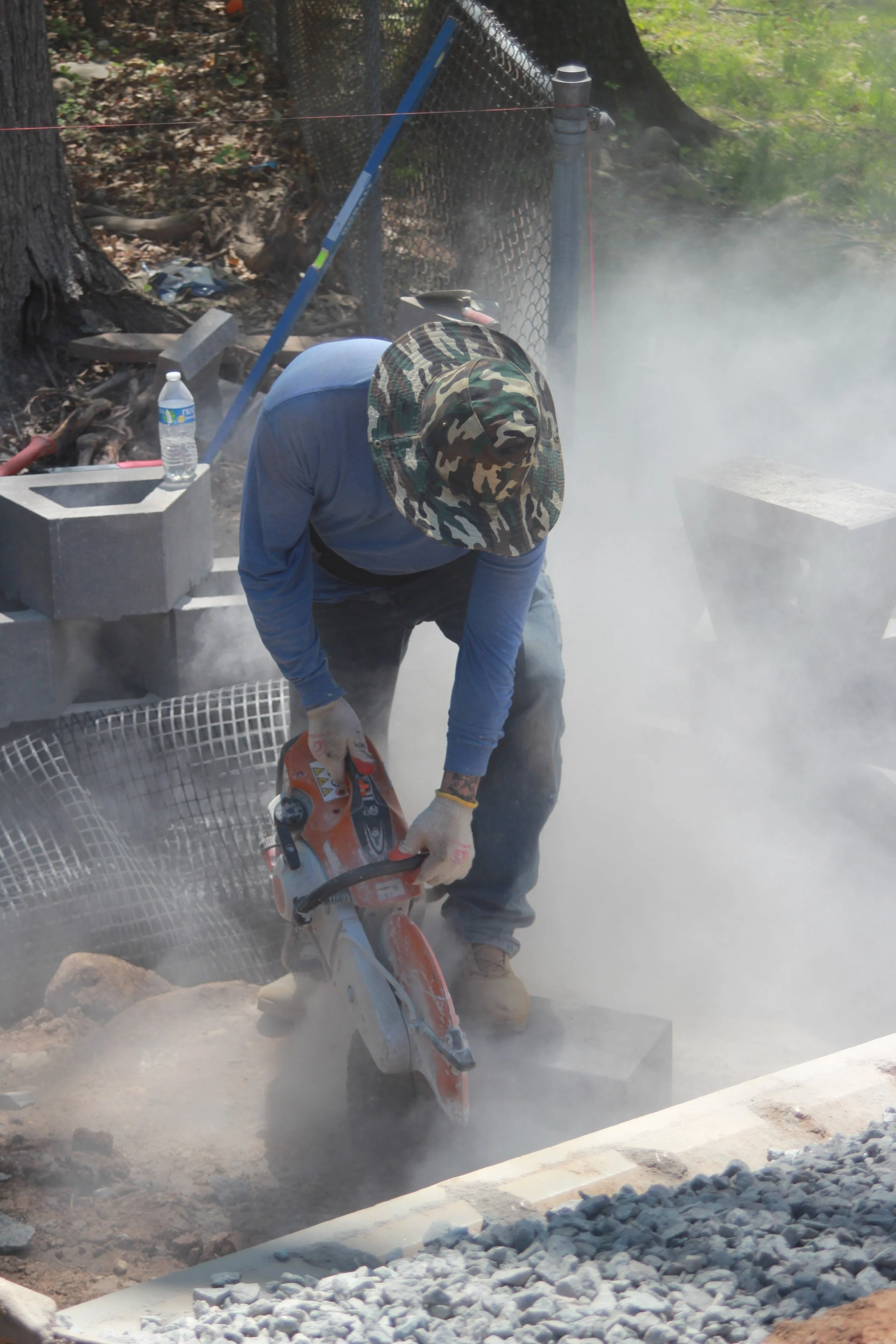 A person wearing a camouflage hat, blue long sleeve shirt, and gloves is cutting concrete with a small orange power saw, surrounded by dust and smoke.