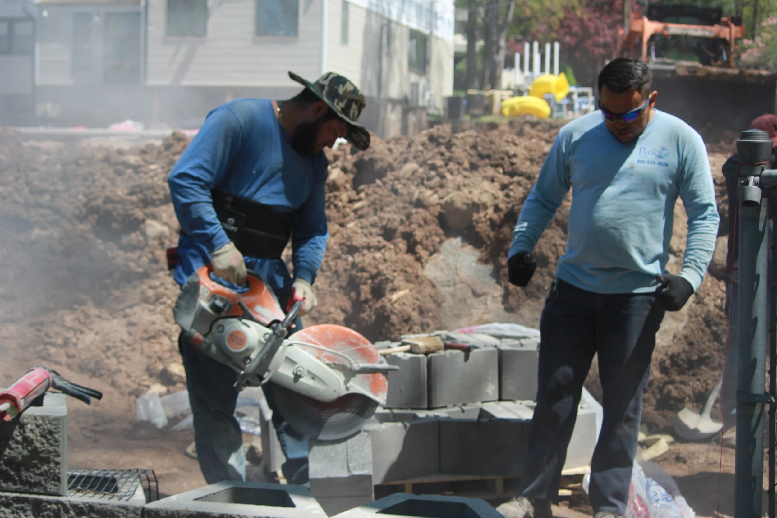 Two construction workers building a brick wall at a construction site, with one using a masonry saw and the other inspecting bricks.