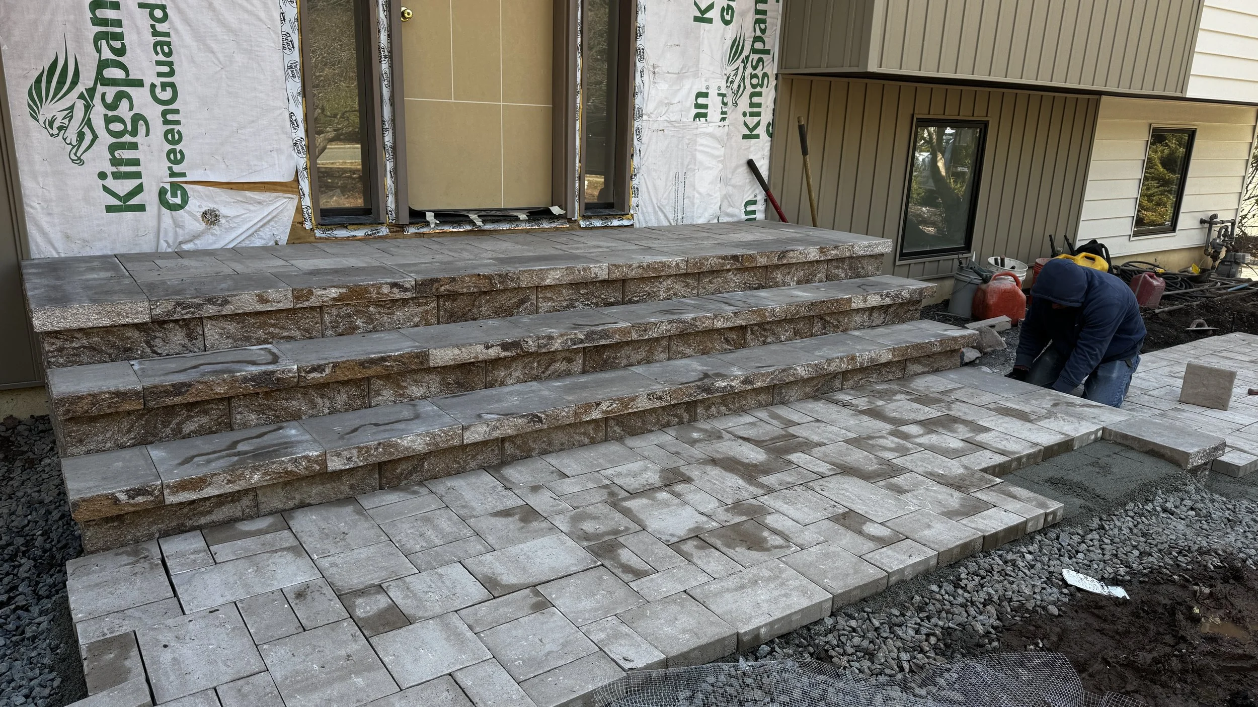 Construction worker installing stone pavers and building steps on a patio outside a house under renovation.