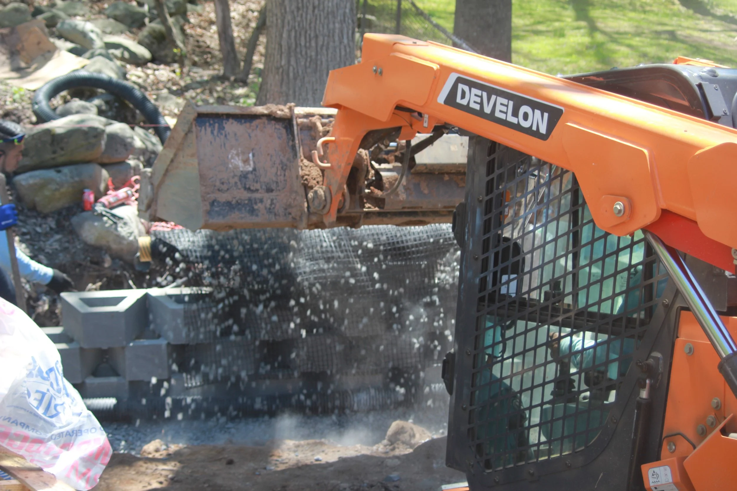 A person operating an orange Develon compact excavator digging into a rocky area, with dirt and rocks falling from the bucket.
