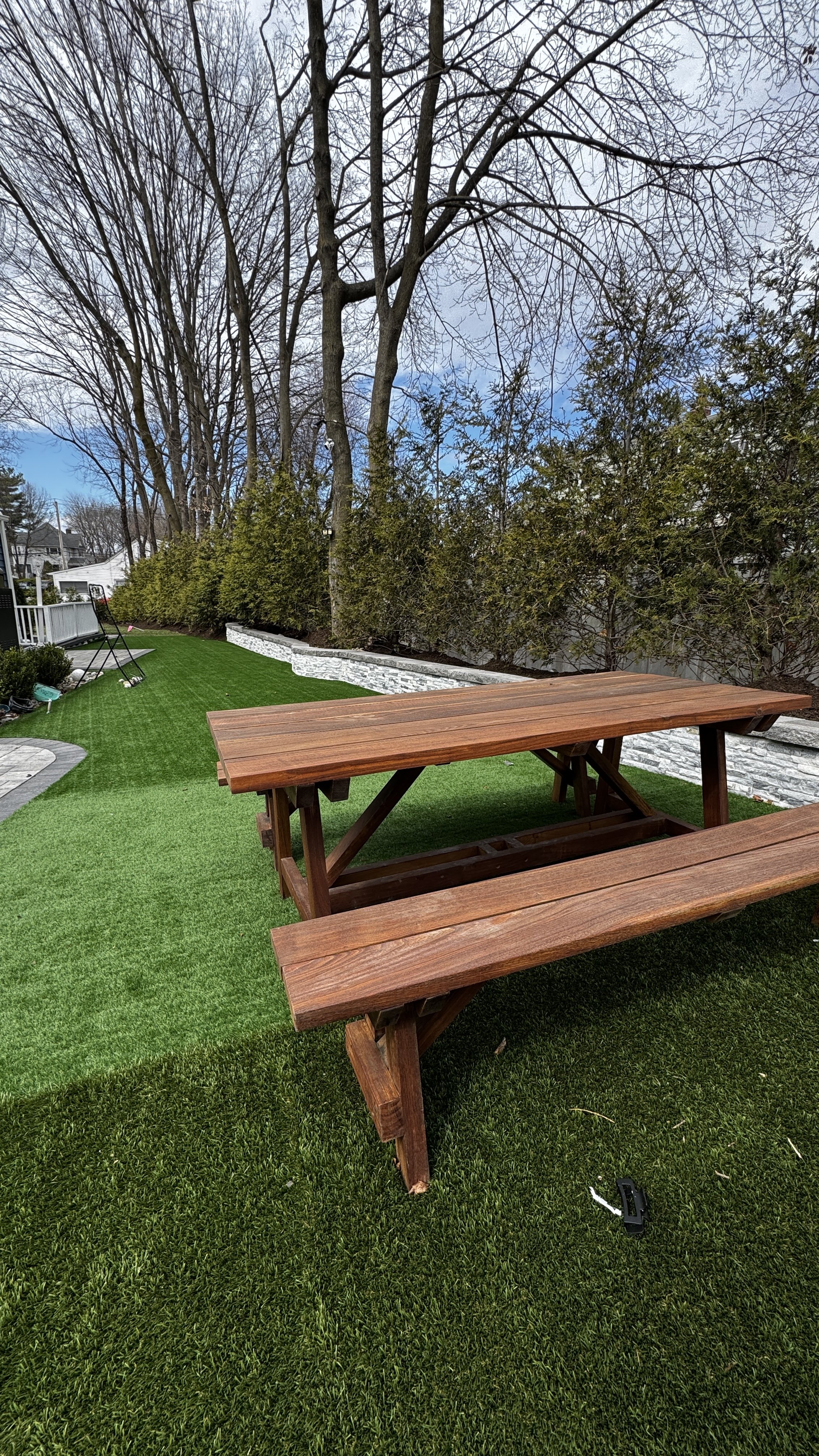 Backyard with a wooden picnic table and green grass, alongside a white brick wall and trees with bare branches.