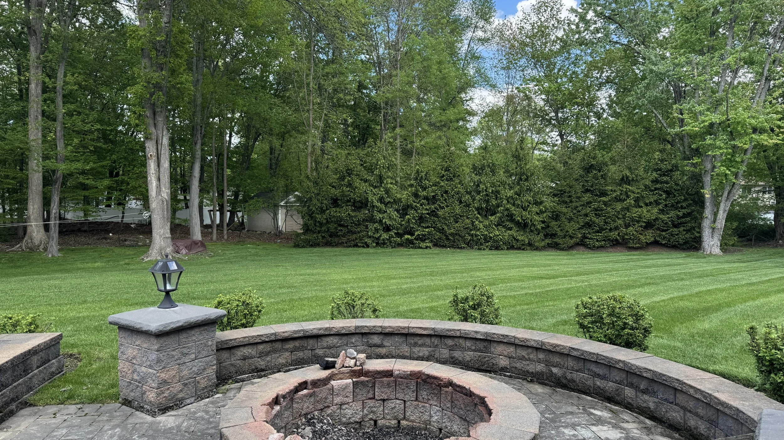 Backyard with a brick fire pit, green lawn, small bushes, mature trees, and shrubs along a fence, with a lantern on a low brick column at the edge of a paved patio.