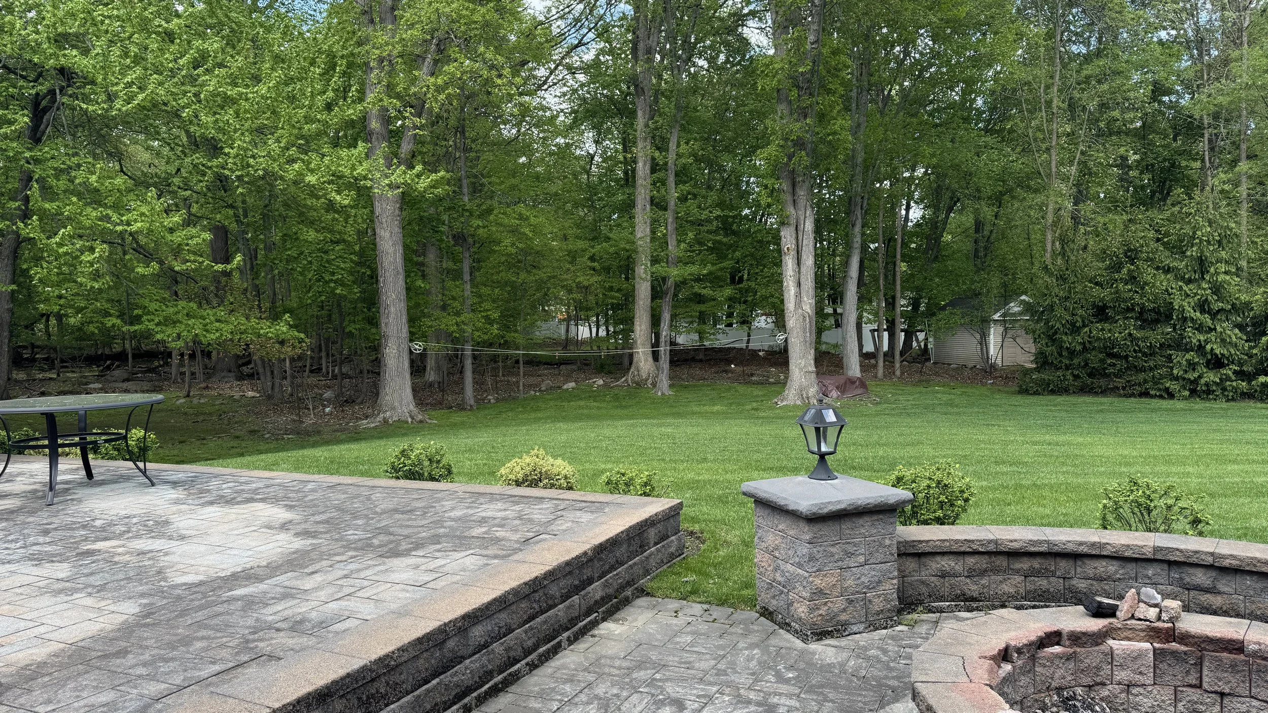 Backyard with patio made of stone pavers, including a small table and chairs, leading to a well-maintained grassy yard with trees and shrubs, and a small shed in the background.