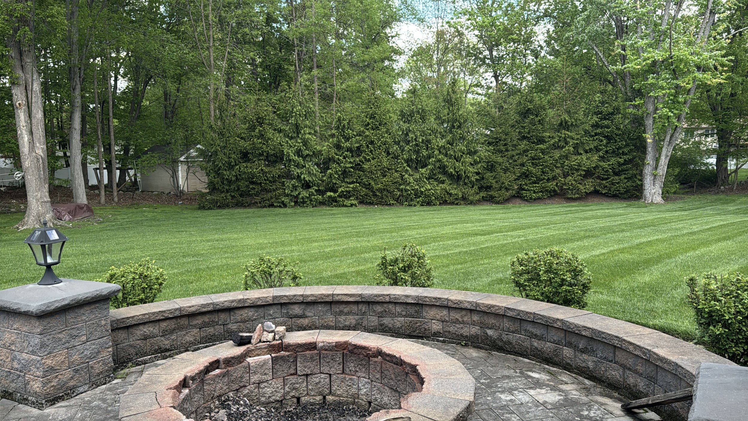Backyard with green grass, trees, shrubs, and a stone fire pit with a brick border in the foreground.