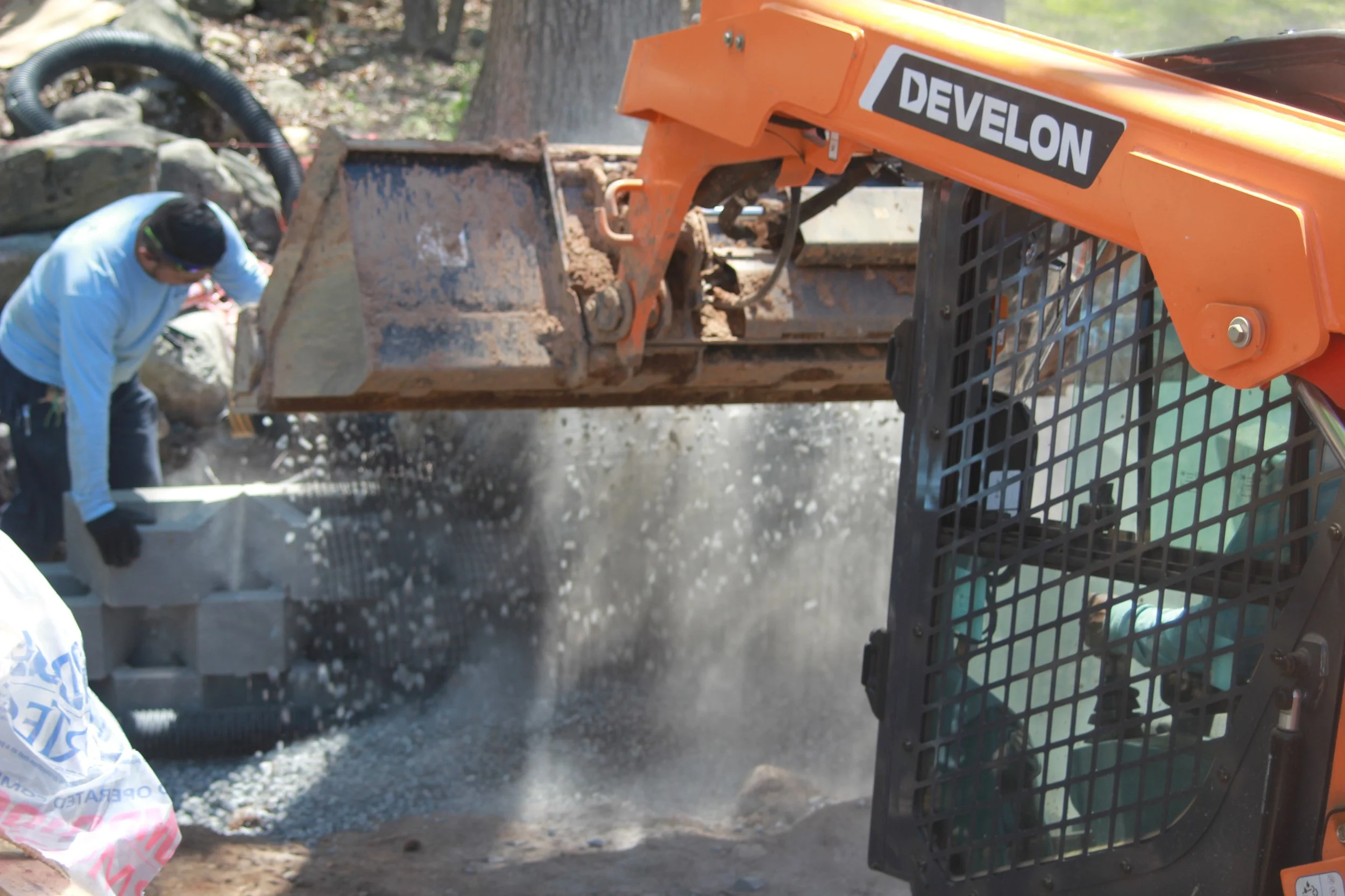 Construction worker operating a mini excavator with an orange arm labeled 'Devlon', lifting and dropping concrete blocks during a construction project.